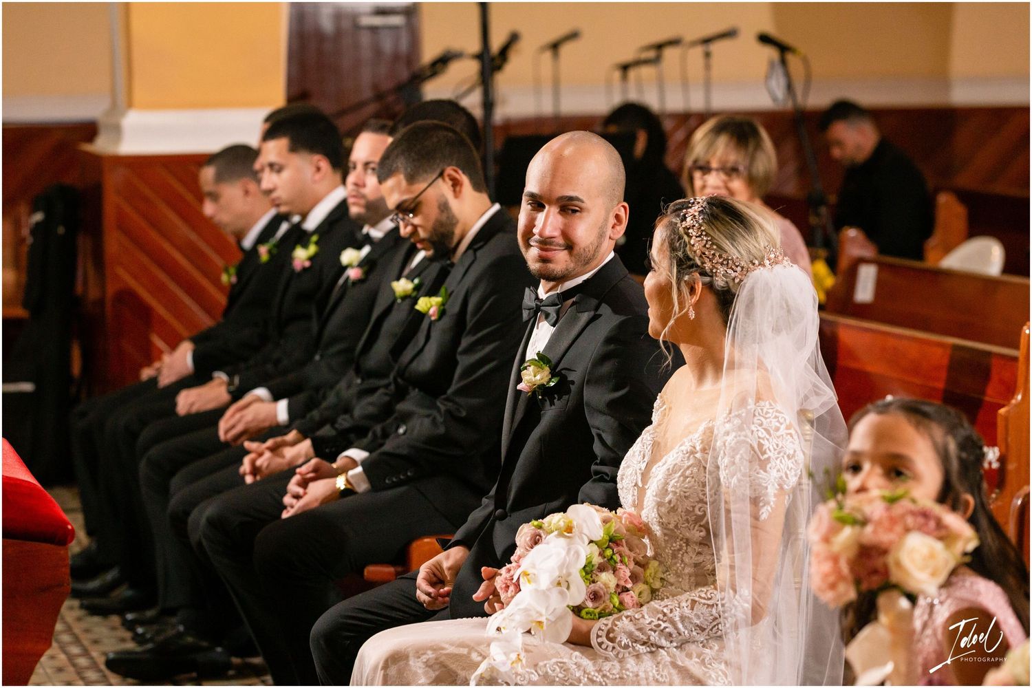 Wedding ceremony participants seated in wooden church pews wearing formal attire.