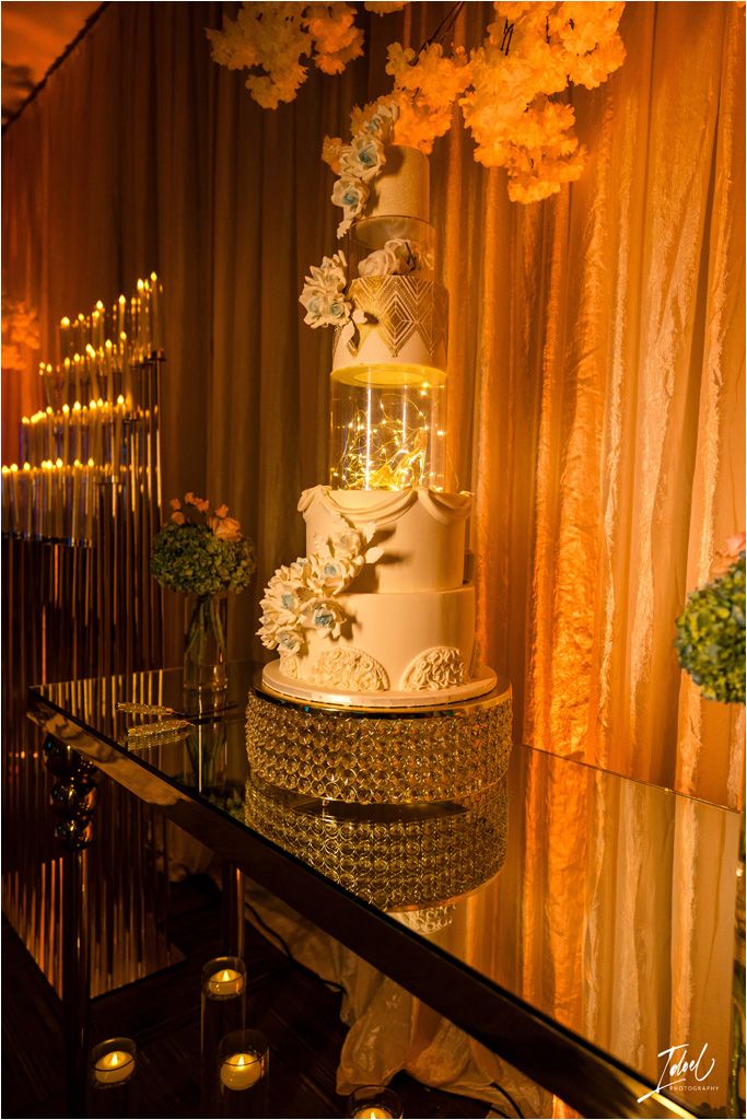 An elegant white tiered wedding cake displayed on a crystal stand with floral decorations and ambient lighting.