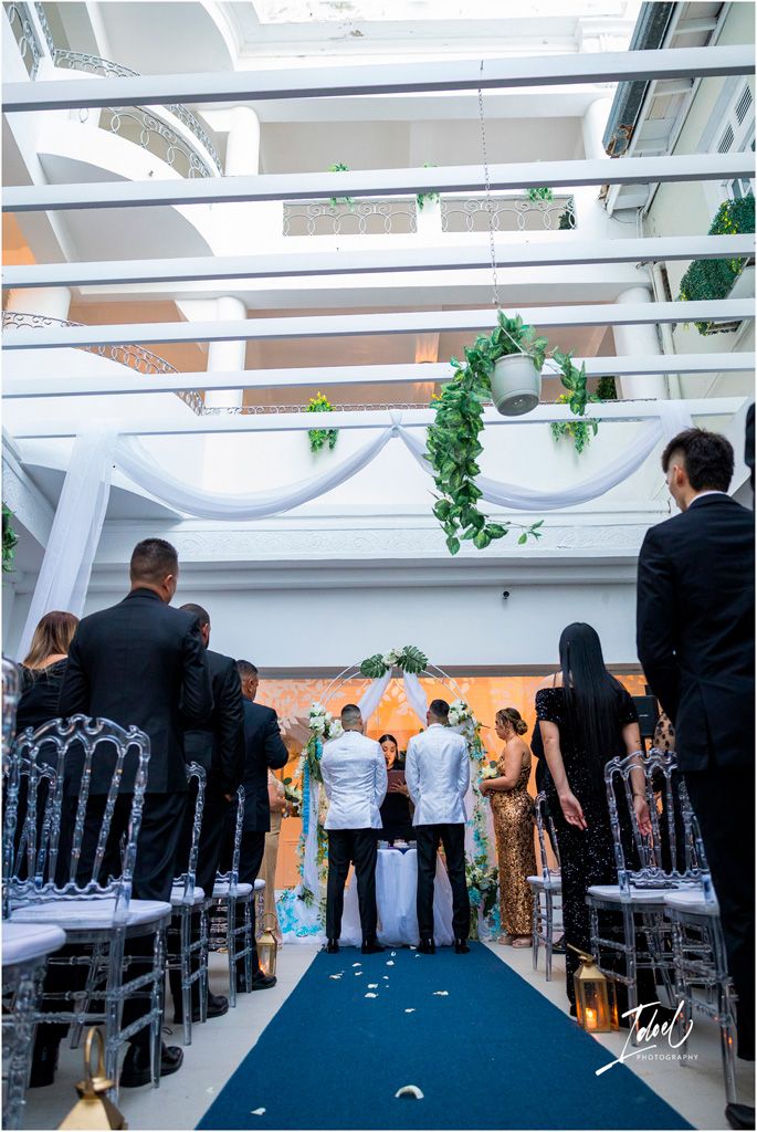 Indoor wedding ceremony setting with white draping, black chairs, blue aisle runner and greenery hanging from ceiling beams.