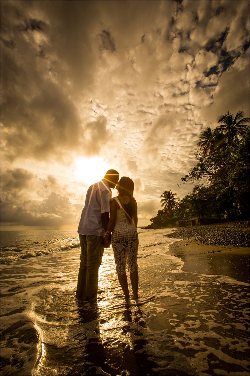A romantic silhouette of a couple standing in shallow ocean water at sunset with dramatic clouds overhead.
