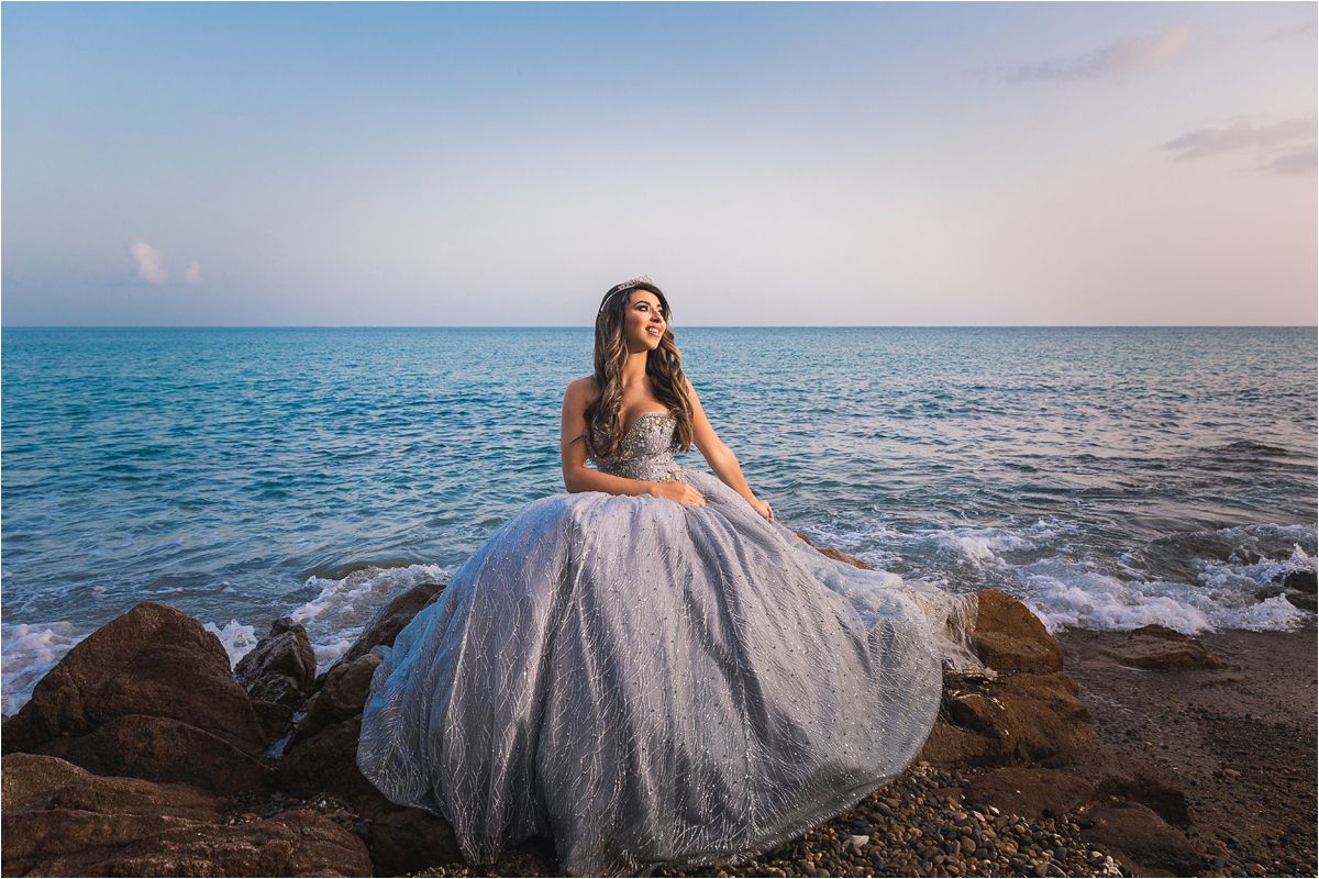 A person in a flowing white gown poses gracefully along a rocky beach coastline at sunset.