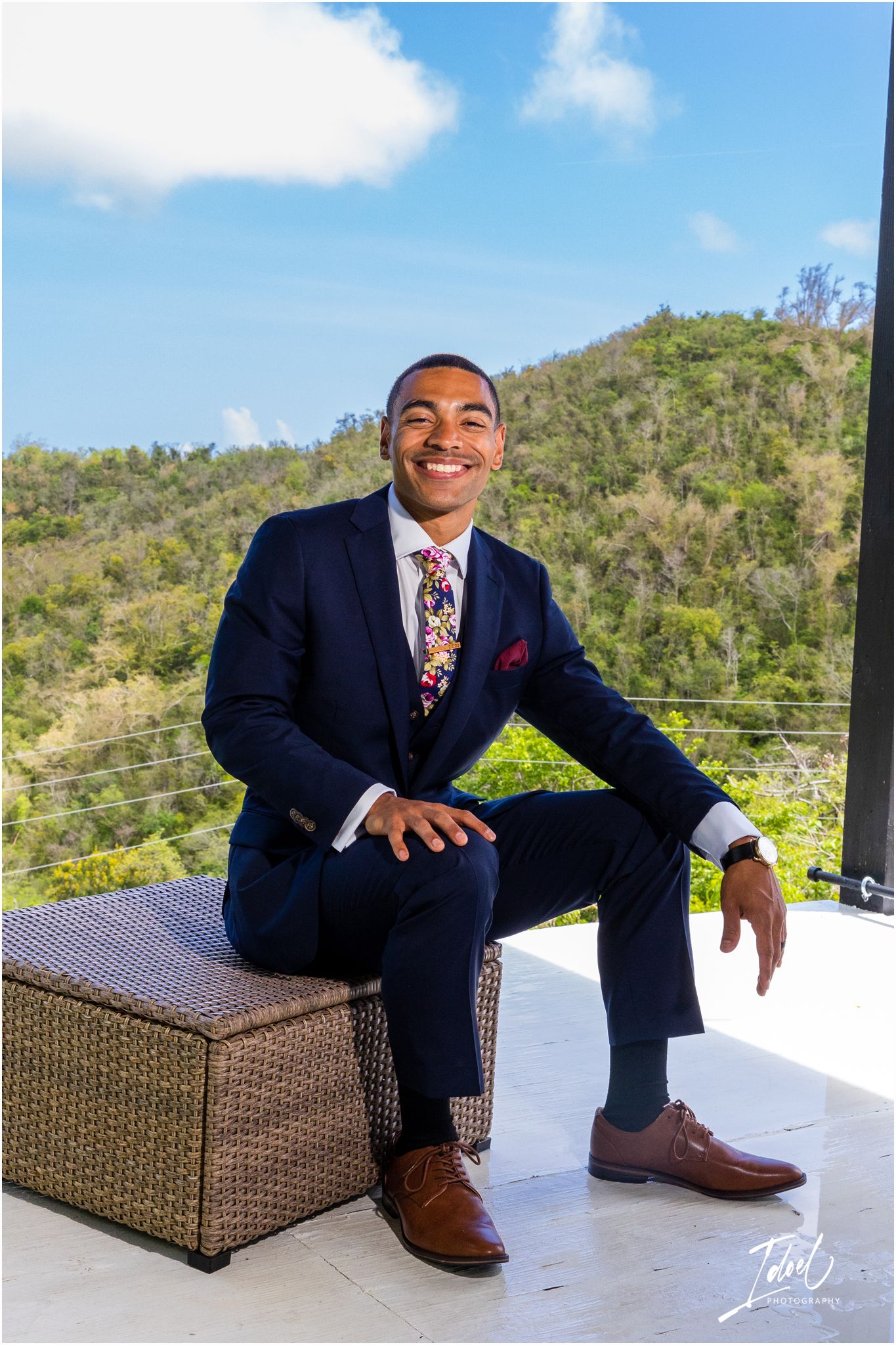 Person in navy suit sits casually on outdoor furniture with mountain view.