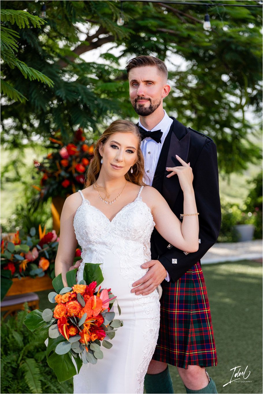 Elegant garden wedding photo with bride in fitted lace dress and groom wearing traditional Scottish kilt.