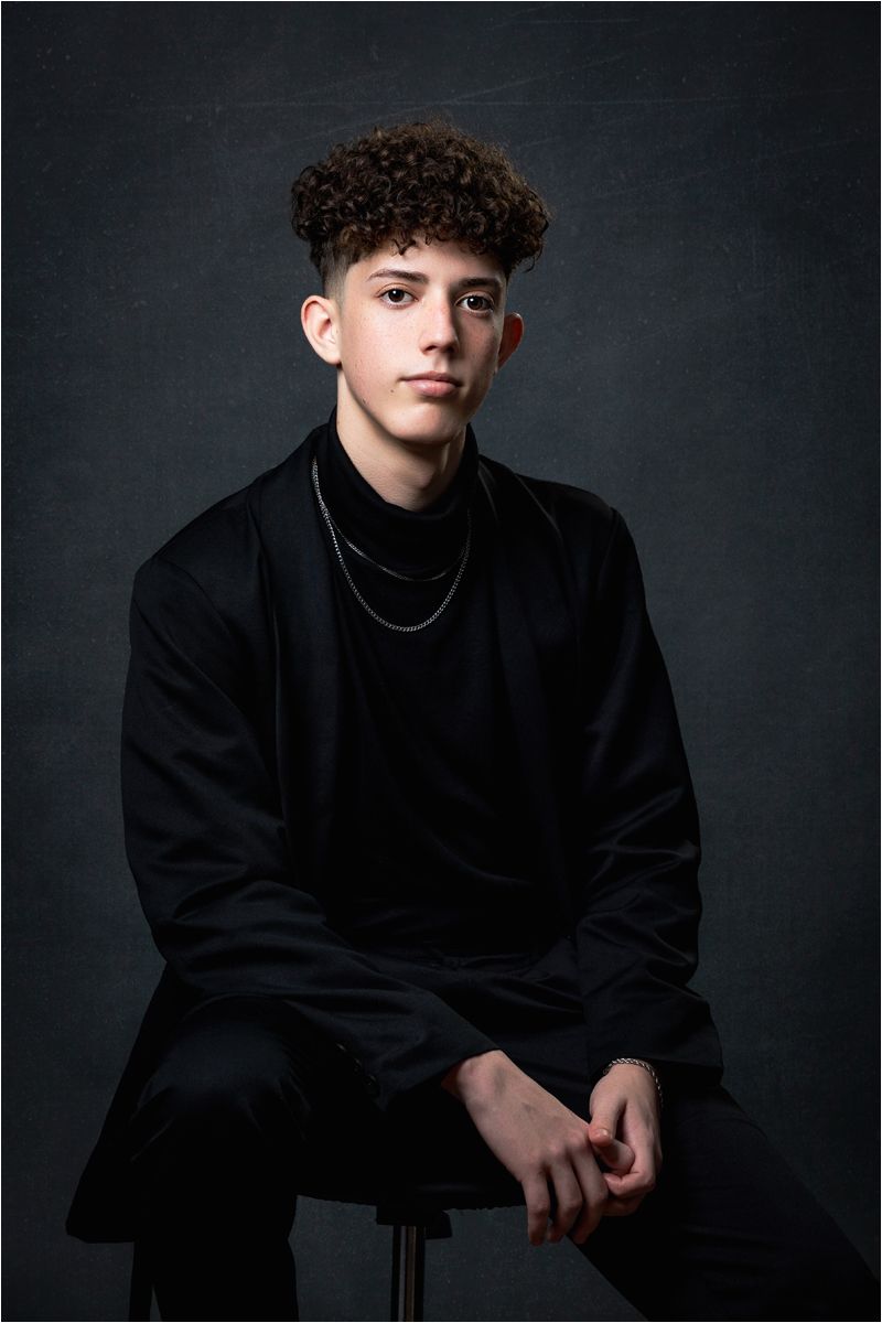 Studio portrait of a young person in all black clothing with curly hair and chain necklace against dark background.