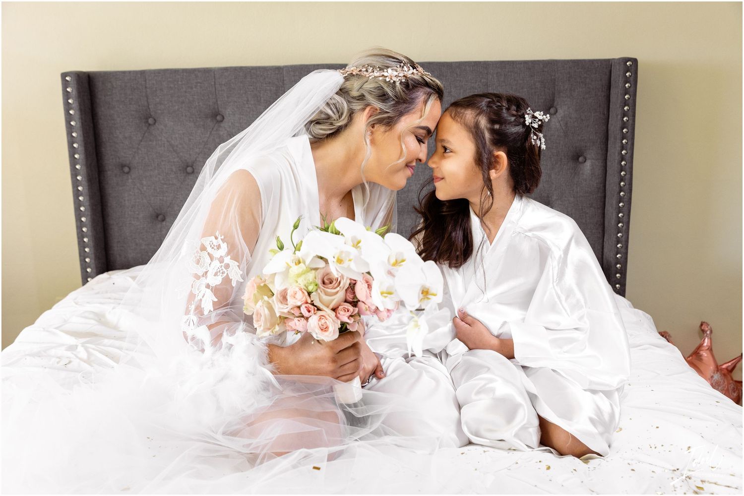 Intimate moment between bride and flower girl in matching white robes on bed with bouquet.