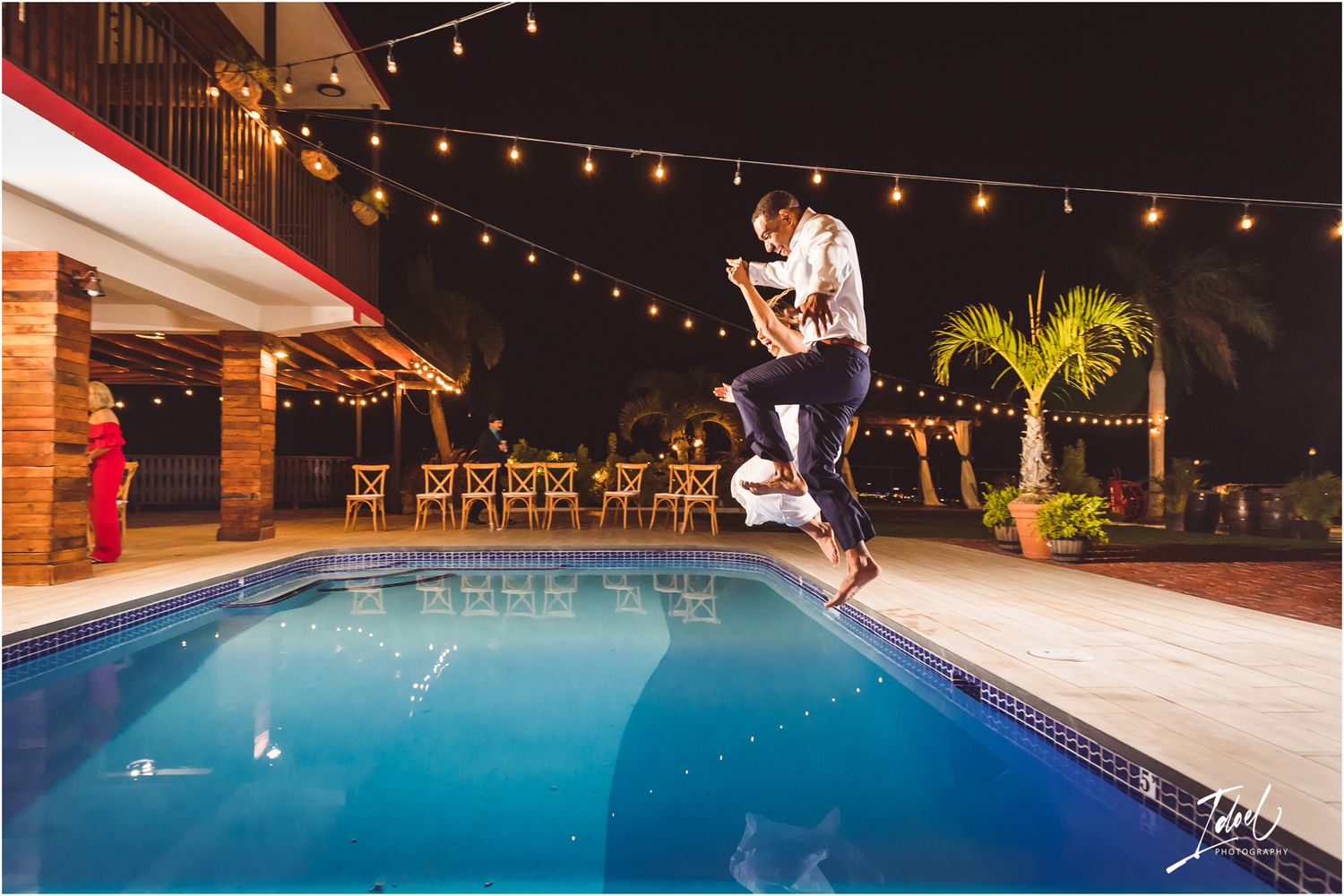 Playful wedding celebration moment jumping into a lit swimming pool at night.
