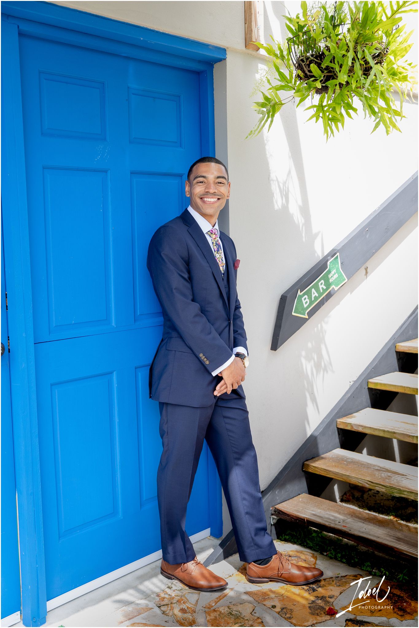 Person in navy suit poses cheerfully by bright blue door and staircase.