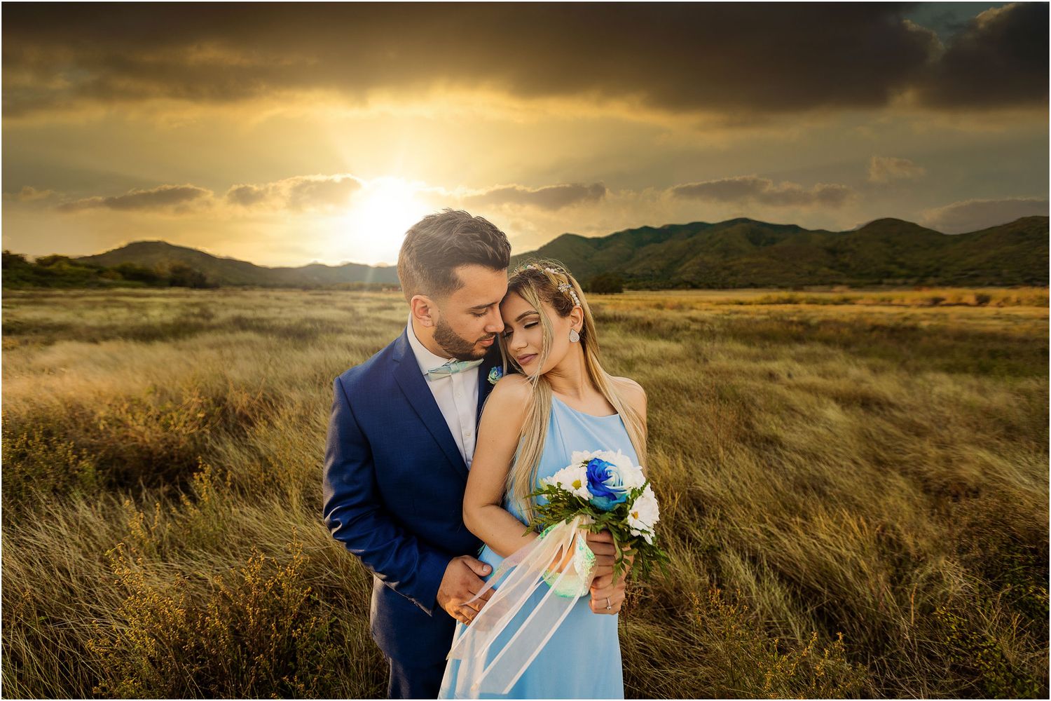 An intimate sunset portrait of a couple in a light blue dress and navy suit amid tall grass.