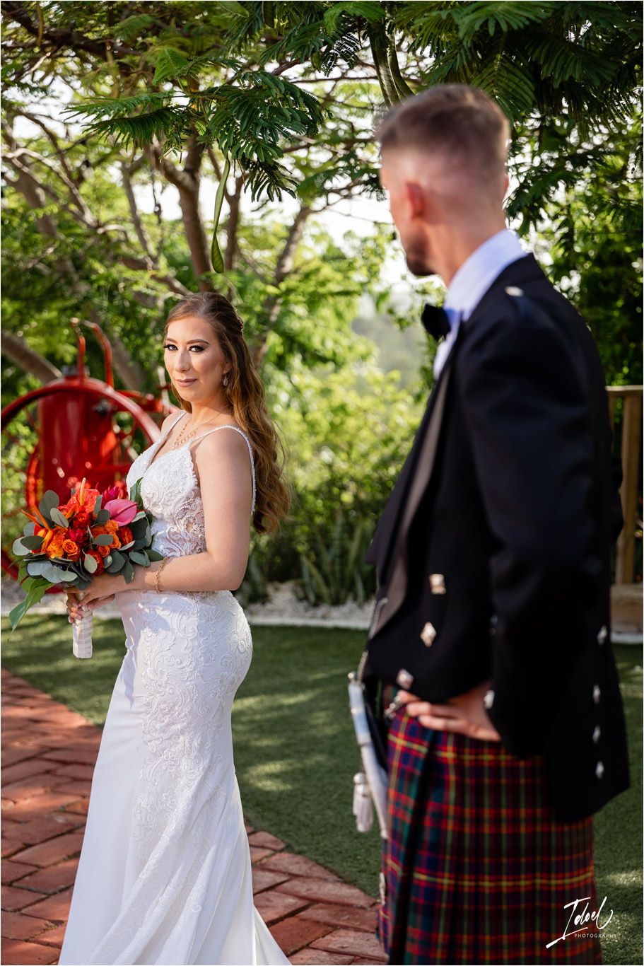 Garden wedding scene with bride holding vibrant coral bouquet and groom wearing traditional Highland dress.