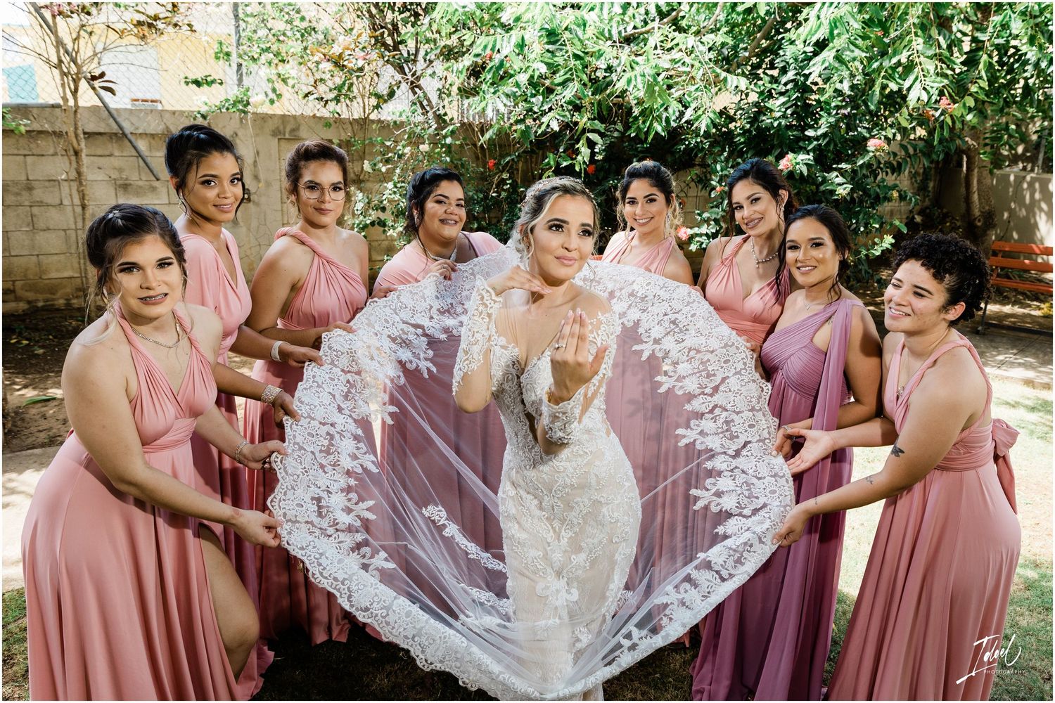 Bridesmaids in pink dresses hold up bride's veil for dramatic outdoor wedding photo.
