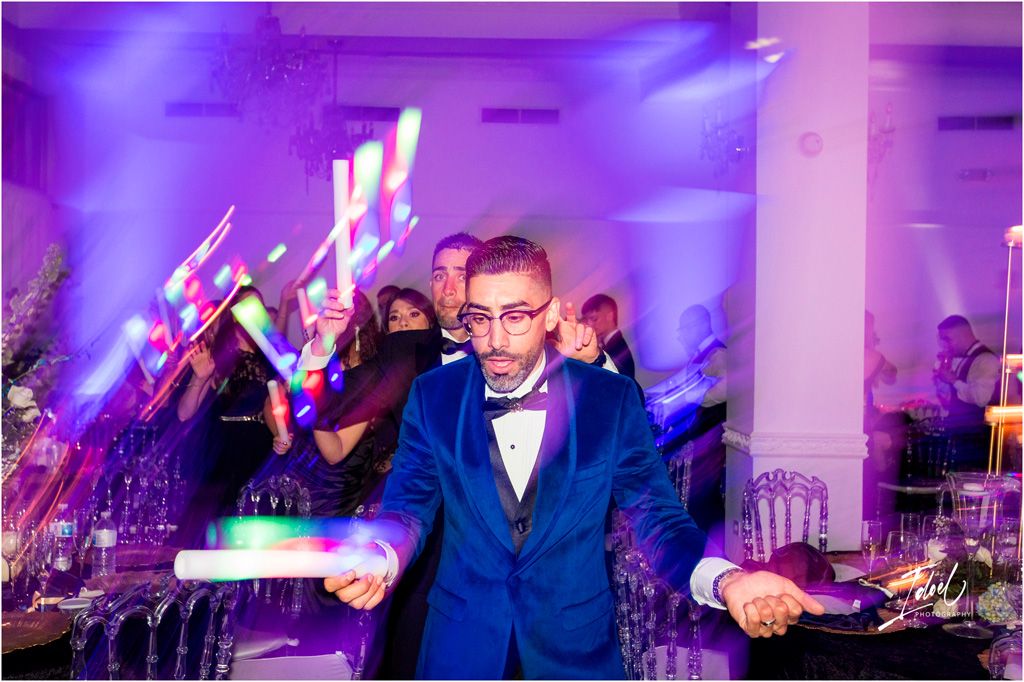 A person in a bright blue suit and bow tie dances enthusiastically at an event with purple lighting and guests in background.