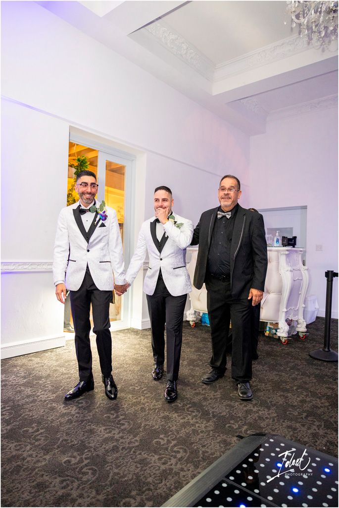 Three people in formal attire stand together at an elegant event venue with white walls and crystal chandeliers.