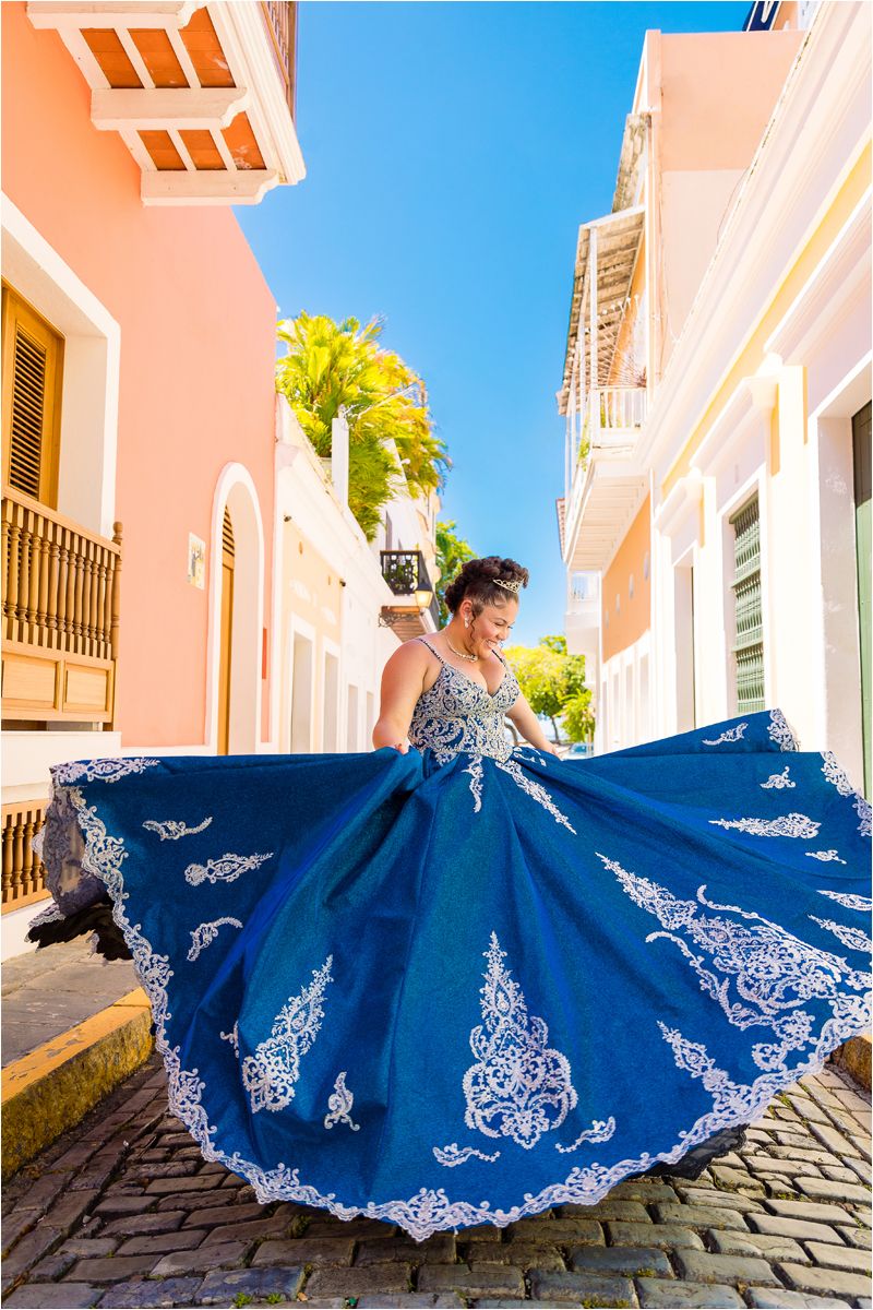 A flowing royal blue dress with white lace embellishments twirls dramatically on cobblestone streets against pink walls.