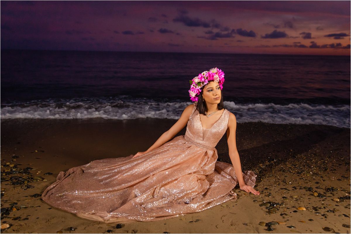 A figure in a flowing pink gown poses gracefully along a beach shoreline at sunset.