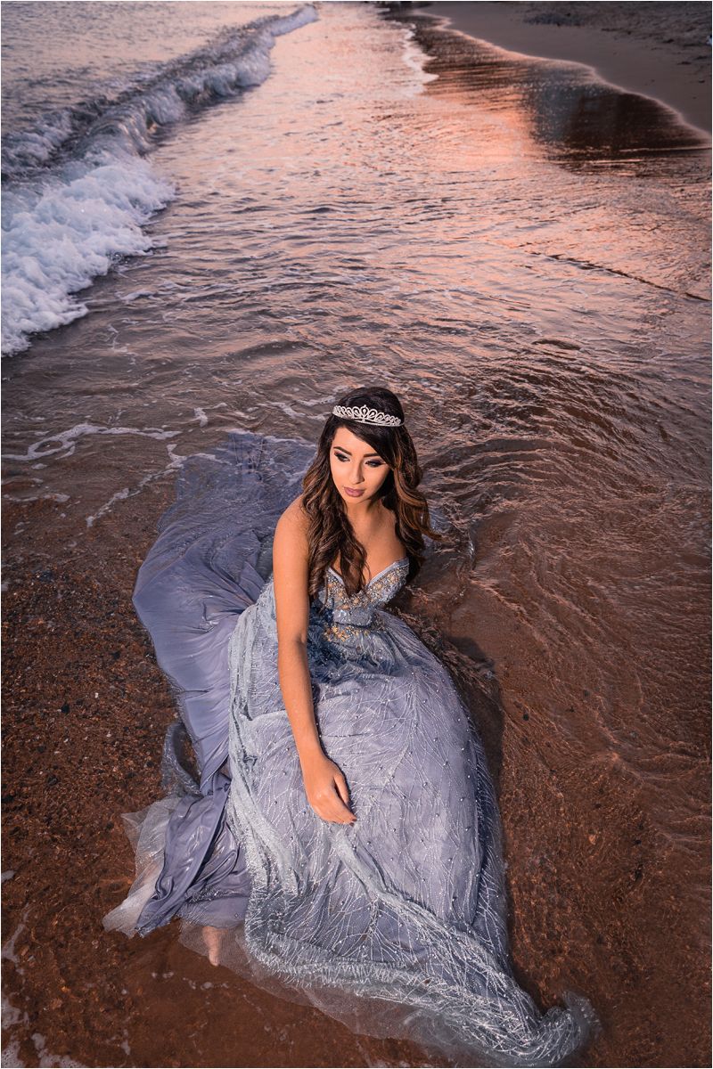 A flowing silver-blue gown sparkles at sunset as waves lap against the sandy beach shore.
