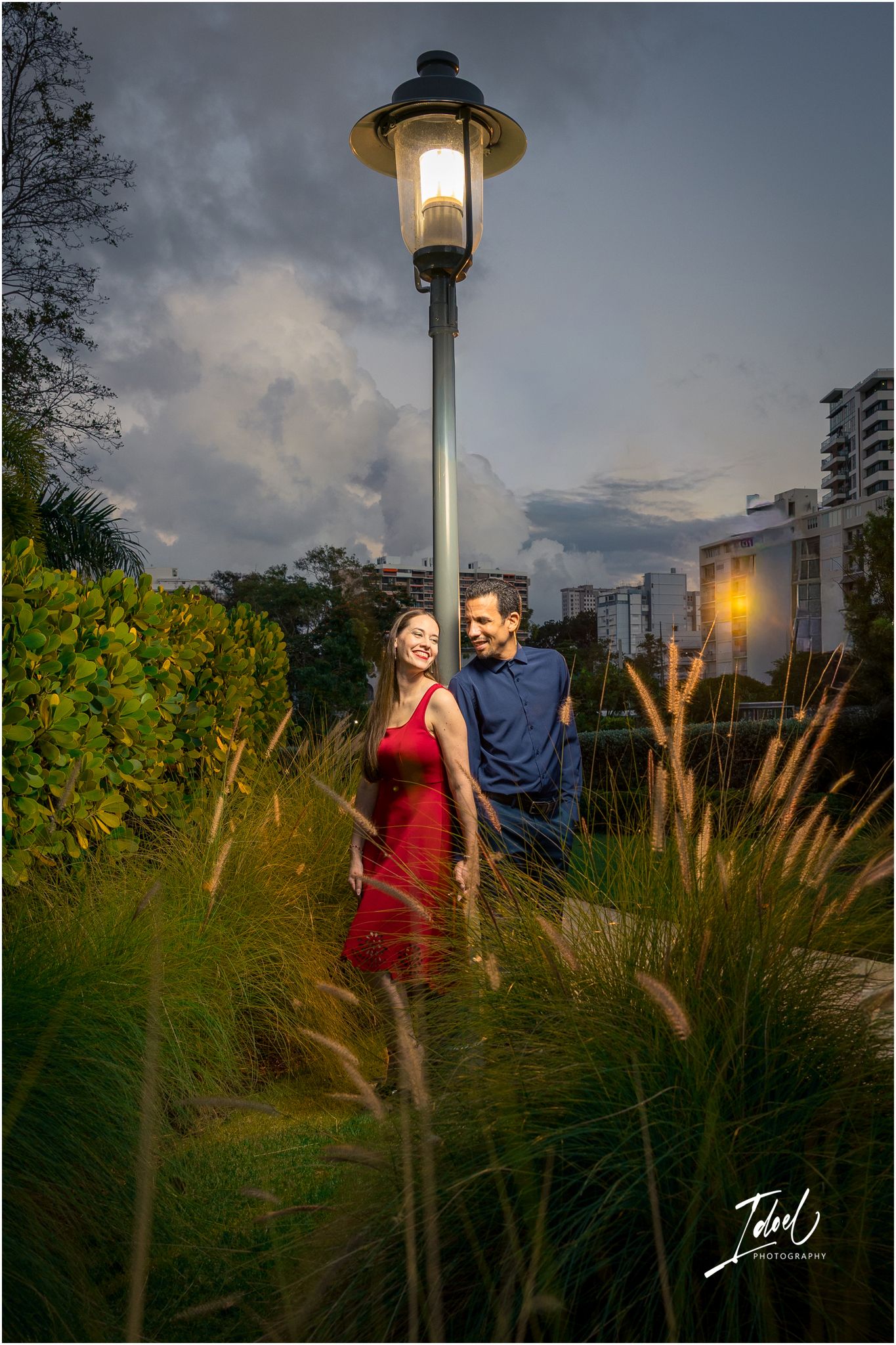 Couple embracing under a glowing street lamp at dusk, surrounded by lush greenery and a dramatic cloudy sky.