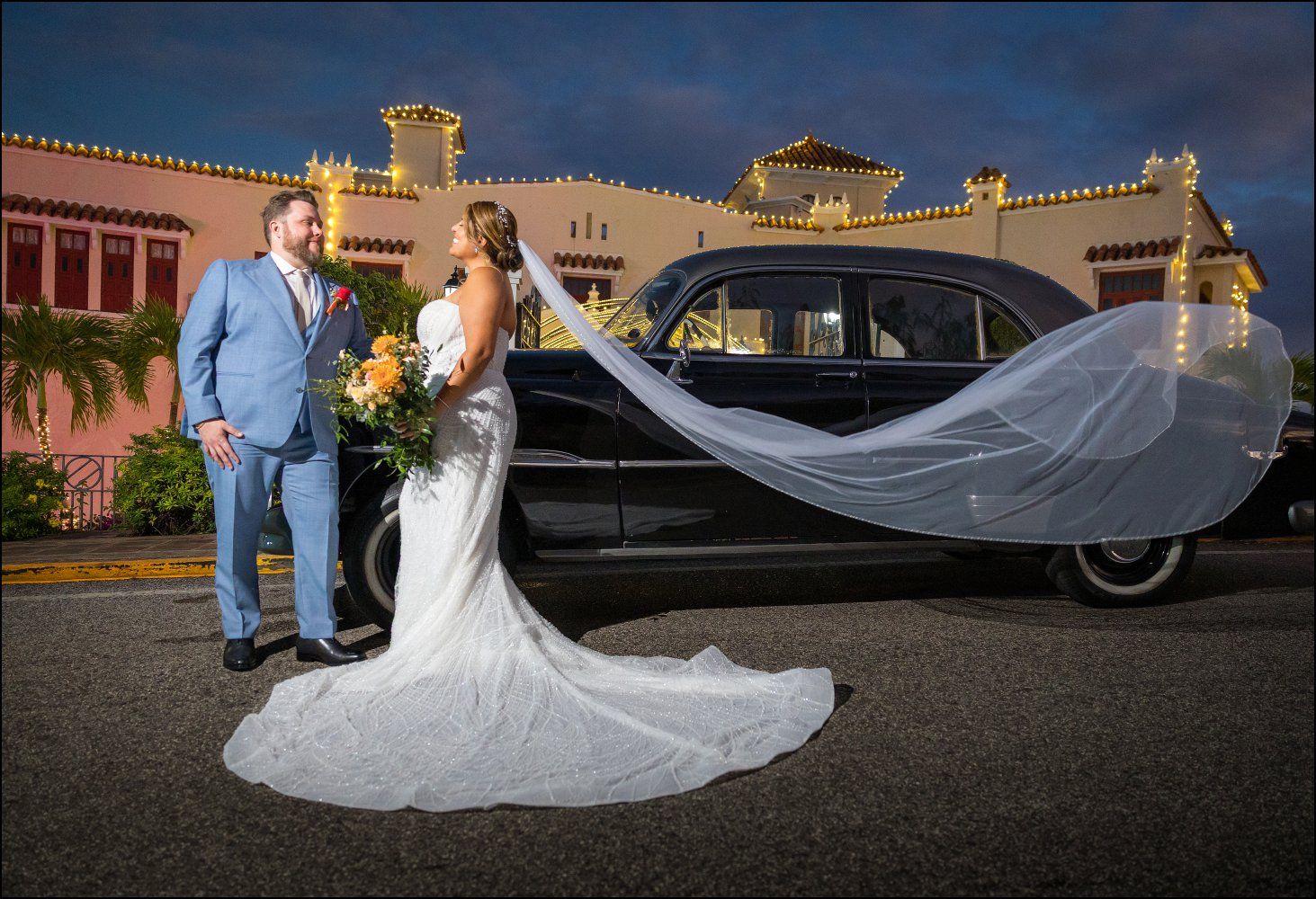 Bride and groom pose by a vintage black car at dusk, with a flowing veil and elegant wedding attire.