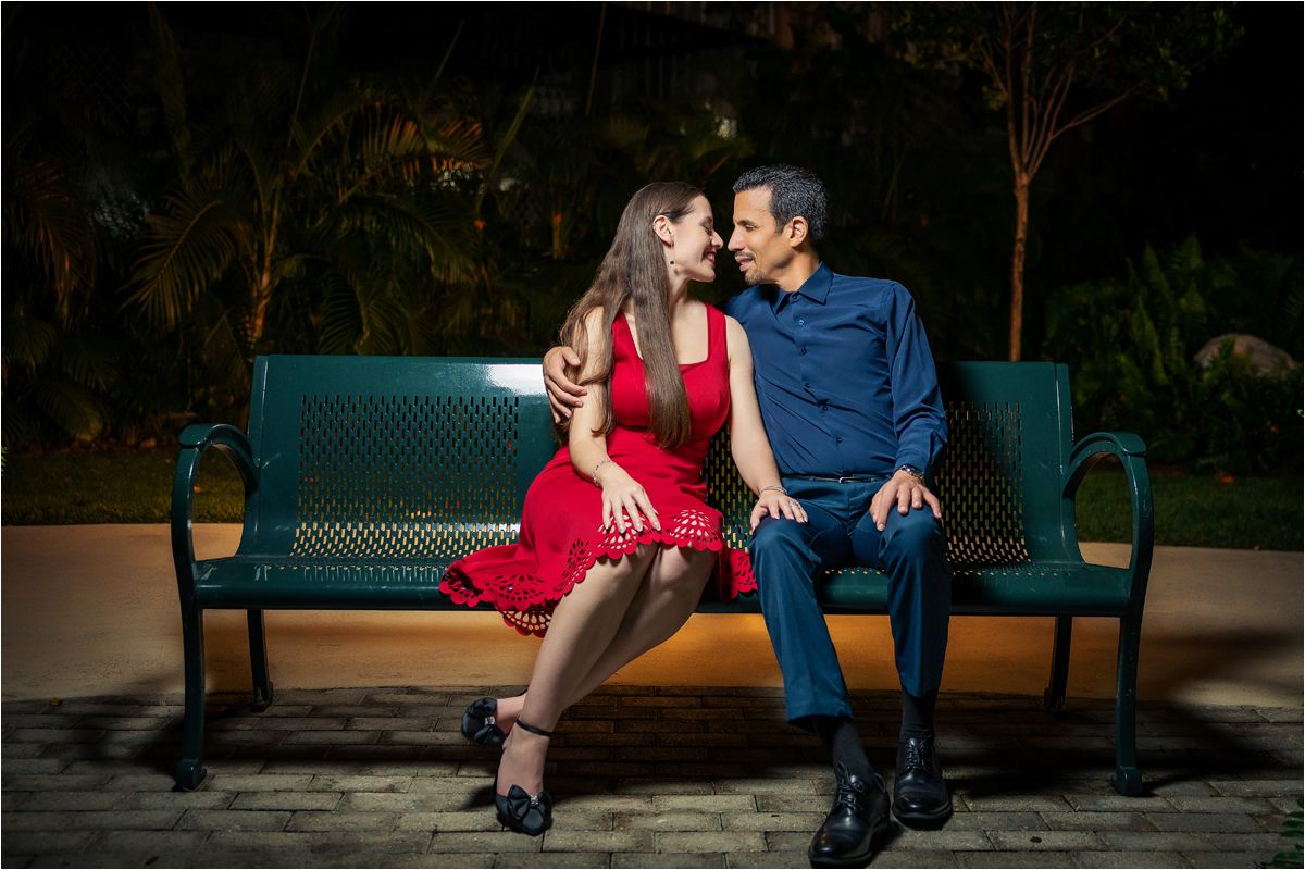 Romantic couple sitting close on a green park bench at night, woman in red dress and man in blue suit.