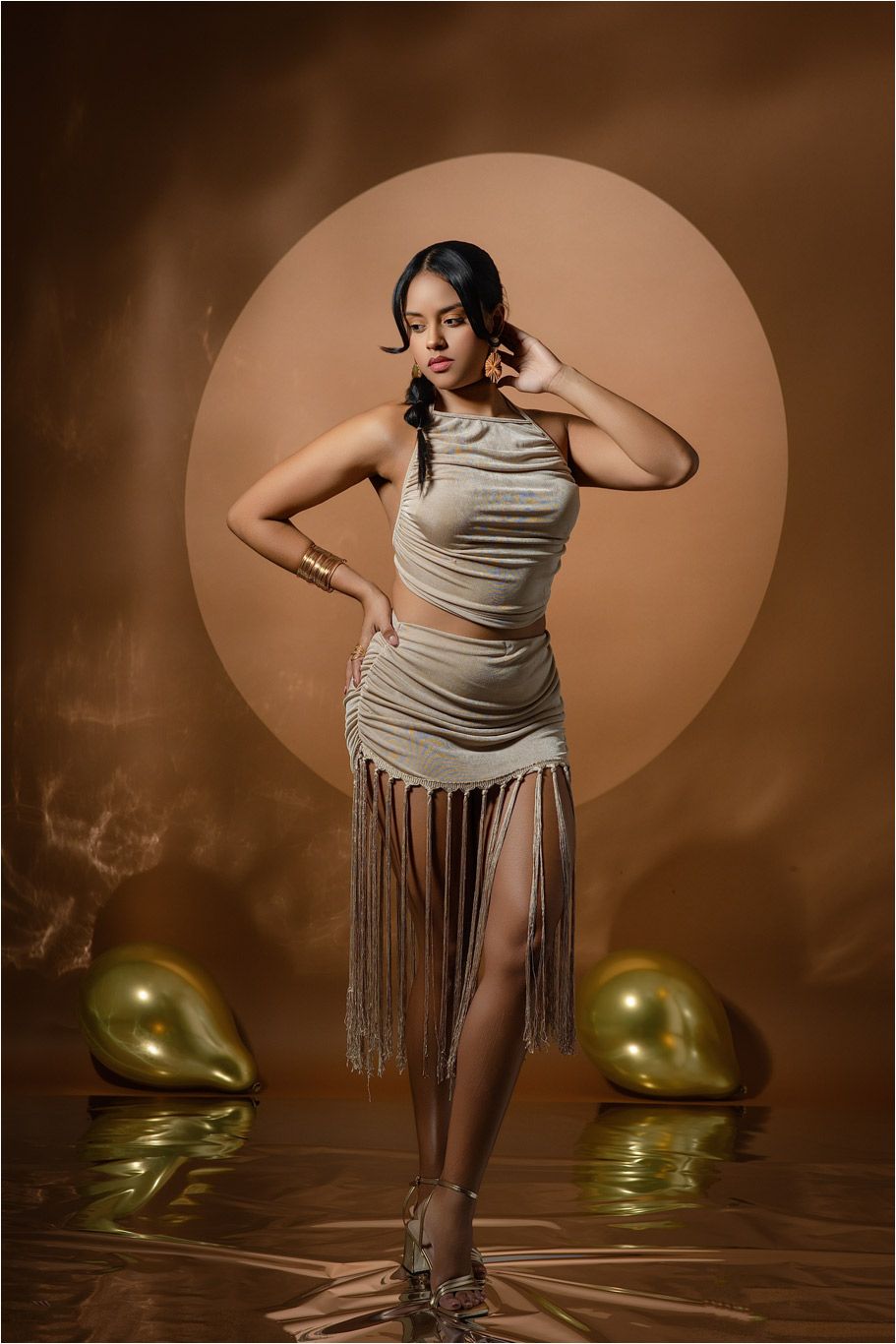 Woman in gold fringe two-piece outfit poses confidently against warm brown backdrop with gold balloons.