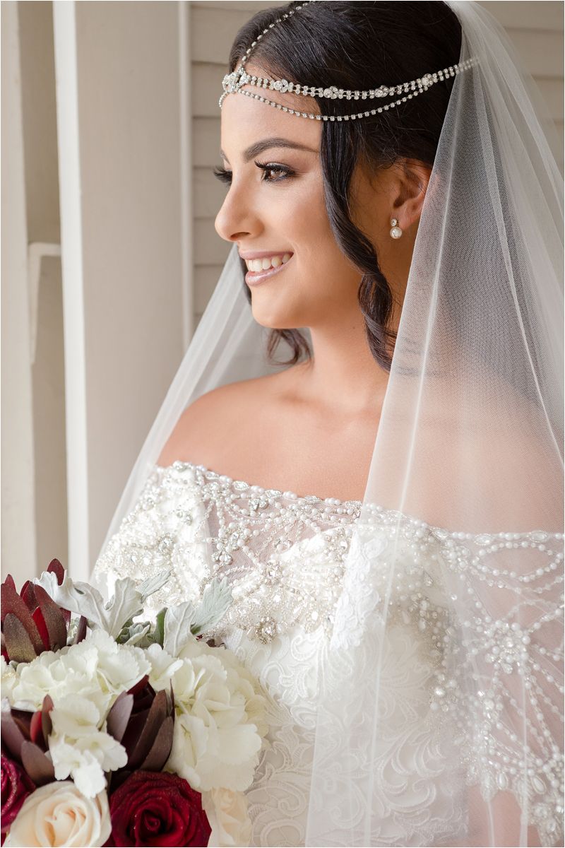 Bride in lace off-shoulder wedding dress wearing crystal headpiece and veil while holding red and white bouquet.