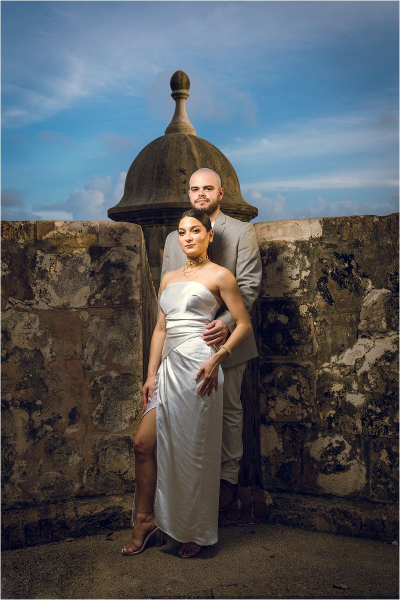A person in a white dress poses dramatically against a stone fortress wall under blue sky.