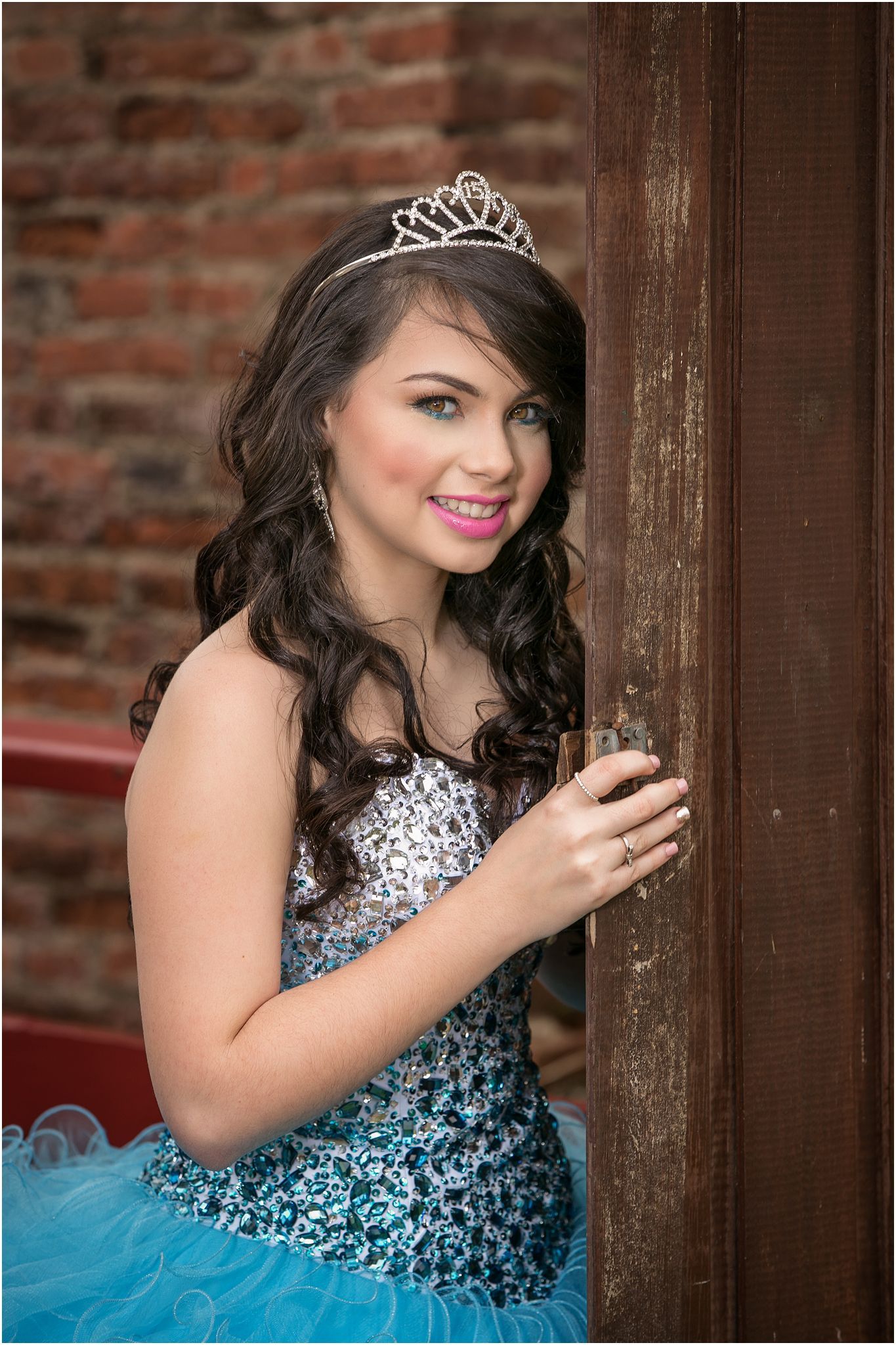 A person in a sparkling blue and silver formal gown with a tiara poses against a rustic brick wall background.