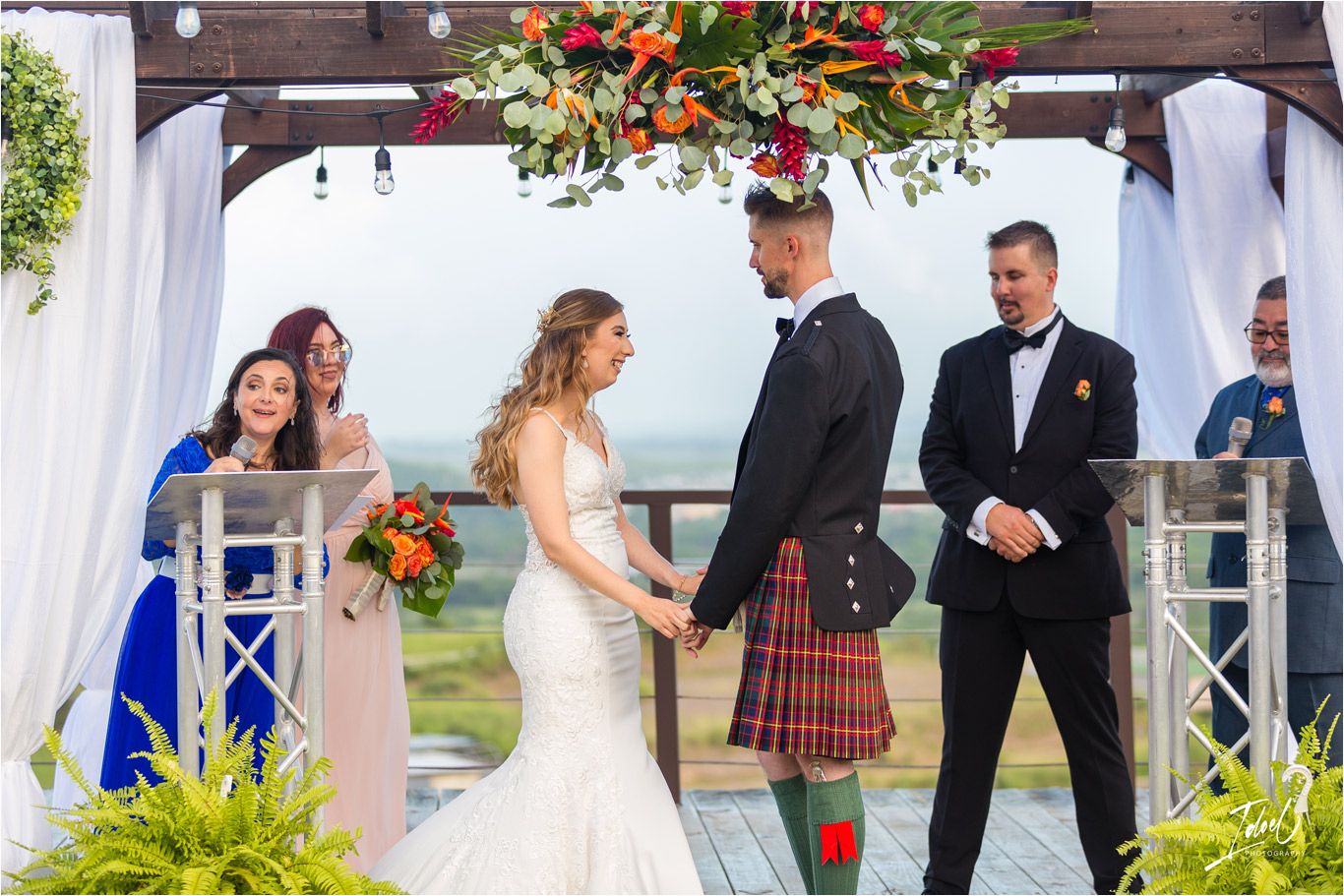 An outdoor wedding ceremony takes place under a white pergola decorated with vibrant flowers and greenery.