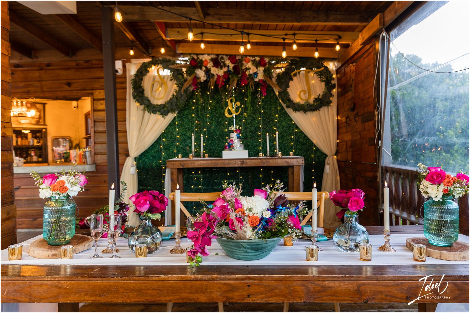 Elegant wedding reception table with pink flowers and greenery arch under rustic wooden beams.