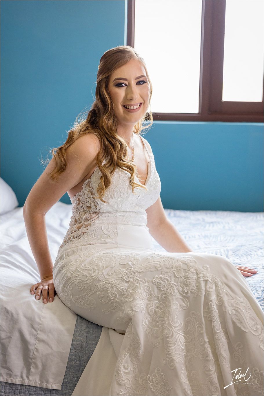 Elegant bride in lace wedding dress sitting on white bed with blue wall background.