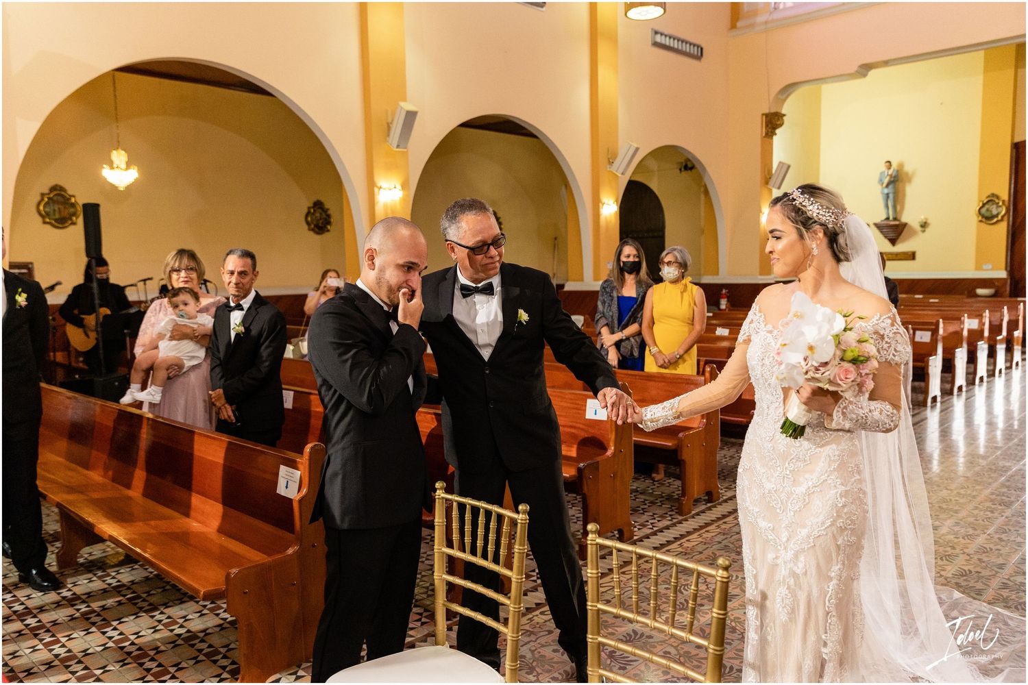 Celebratory scene of wedding guests reacting during ceremony in yellow-walled church with arches.