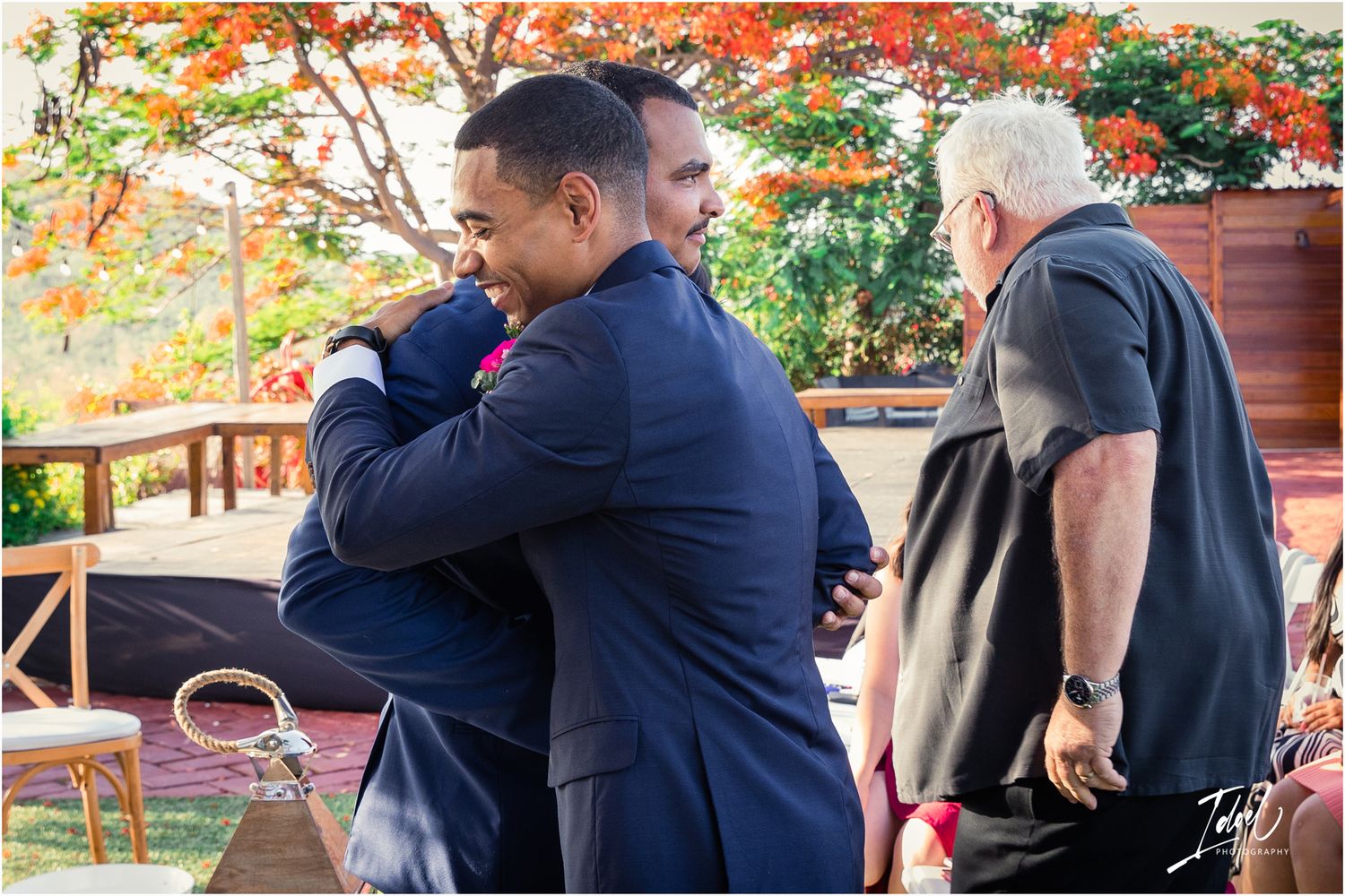 Heartfelt hugs shared between wedding guests during outdoor ceremony with red floral backdrop.