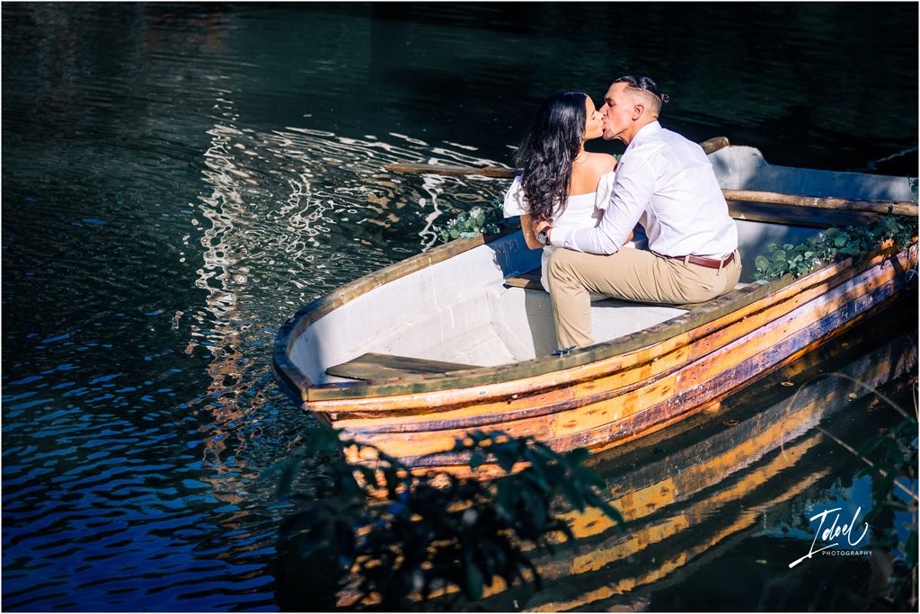 A couple shares a romantic moment in a wooden rowboat on sparkling blue water at dusk.