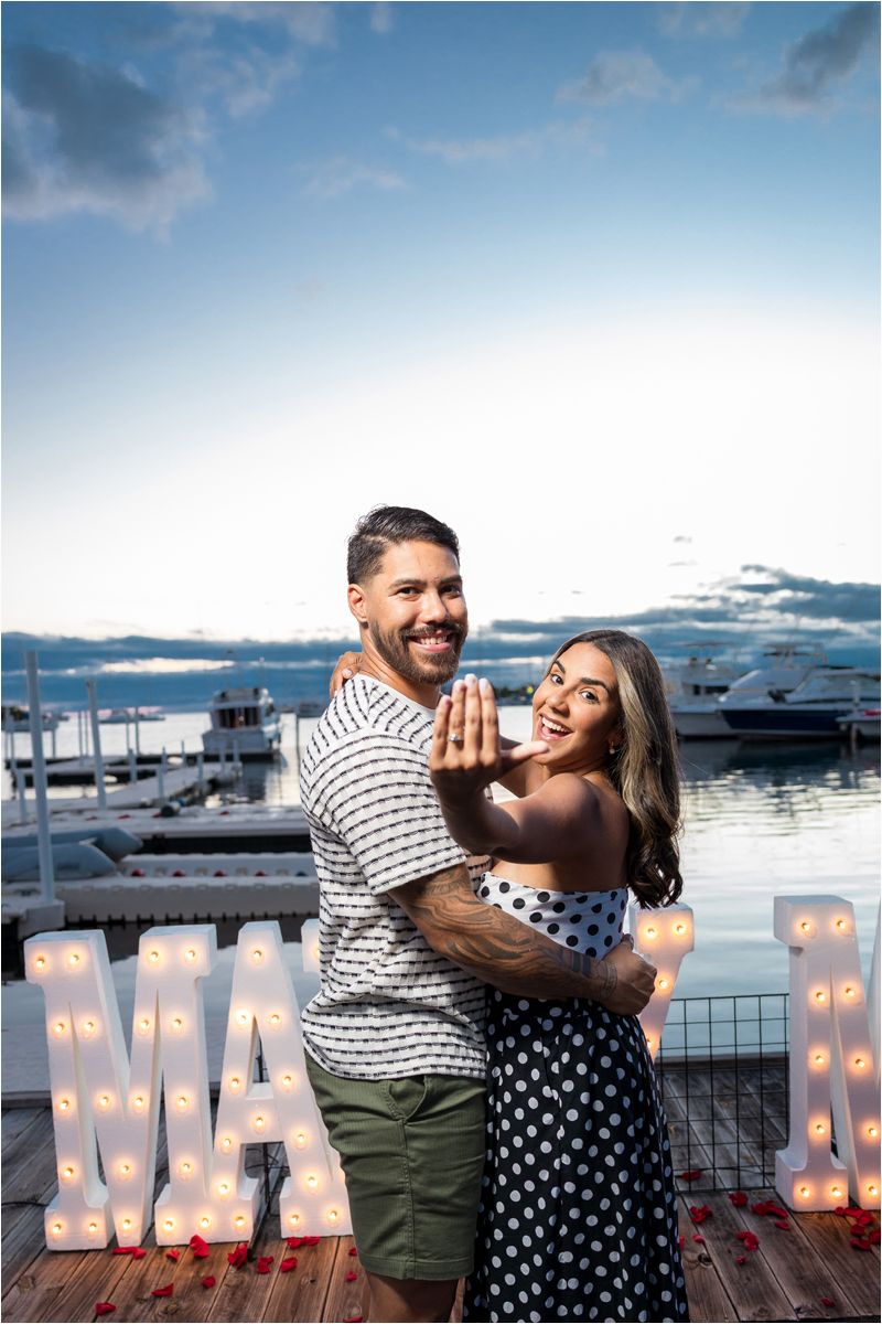 A joyful couple embraces at a marina dock at sunset with illuminated letters and yachts in the background.