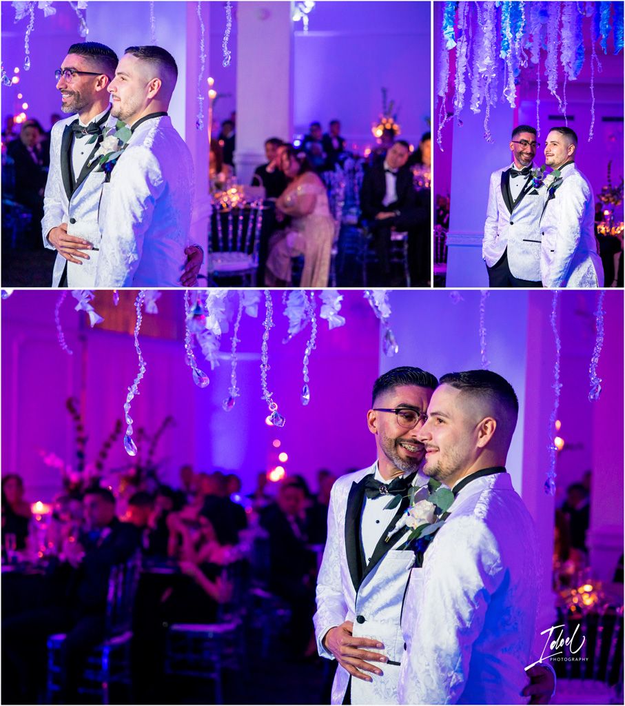 A couple in matching white tuxedos dances under purple lighting and hanging decorations at their elegant wedding reception.