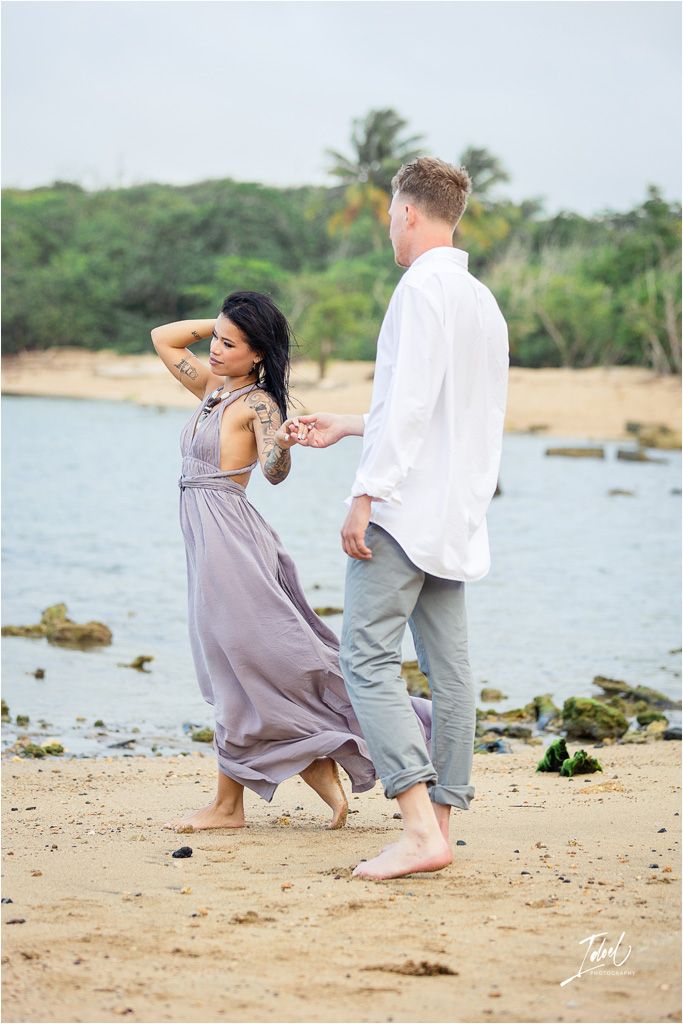 Couple in casual attire dancing barefoot on beach with ocean view.