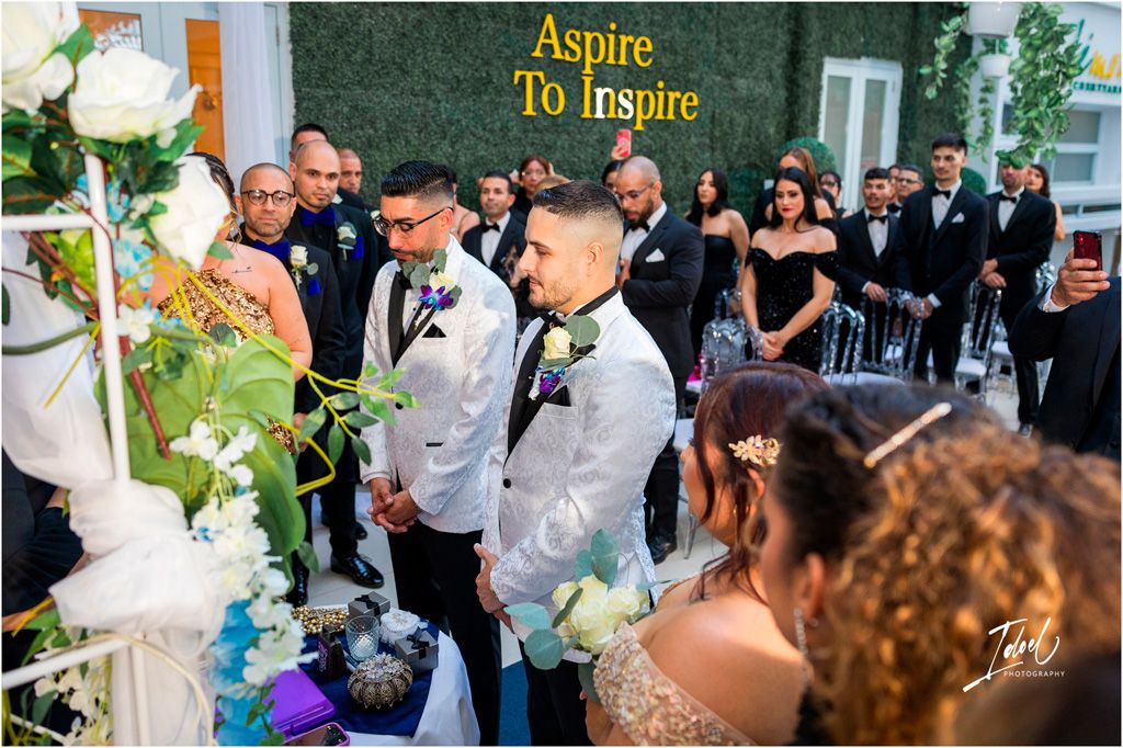 Two people in matching gray suits stand before an altar decorated with white and purple flowers at a wedding ceremony.
