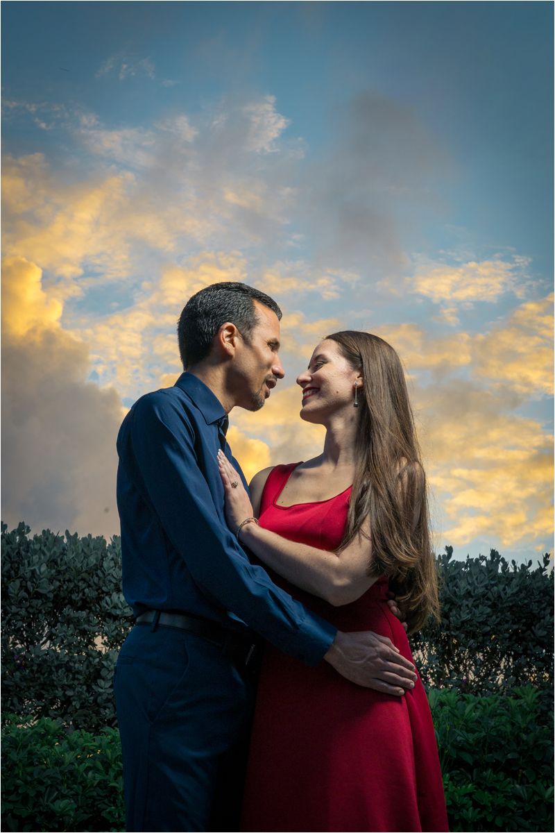 Couple in romantic embrace at sunset, man in blue shirt and woman in red dress, with dramatic cloudy sky backdrop.