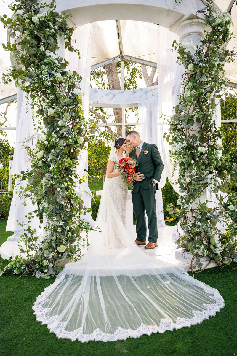 A romantic wedding ceremony scene under a white gazebo decorated with lush greenery and floral arrangements.