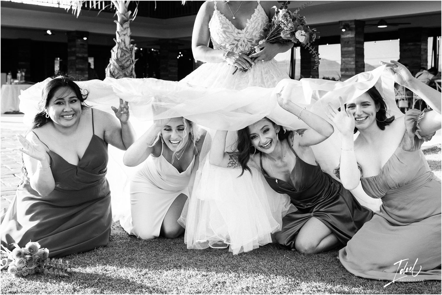 Black and white photo of wedding party peeking out from under bride's tulle dress on lawn.