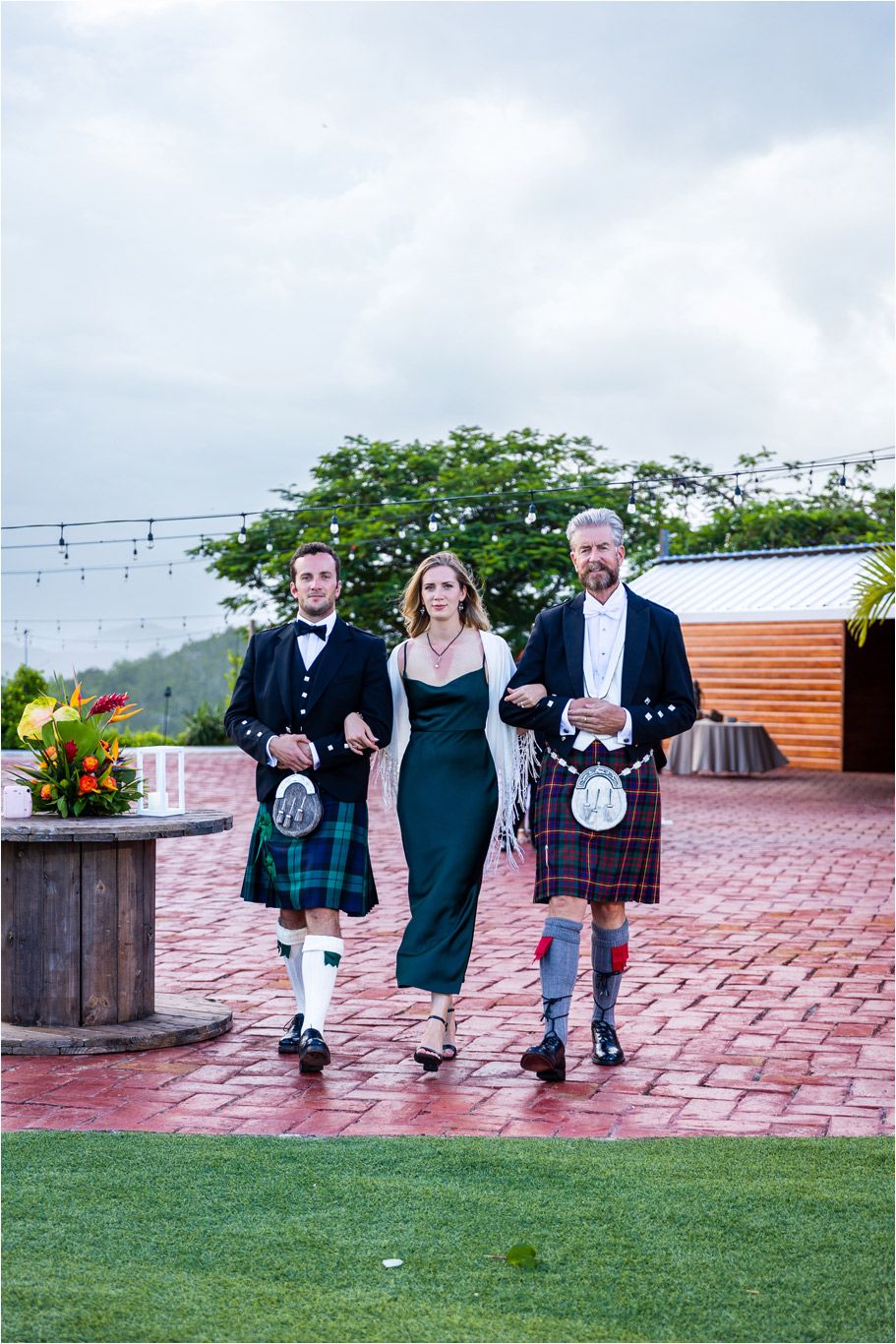 Three people in formal attire including kilts walk together on a brick pathway.