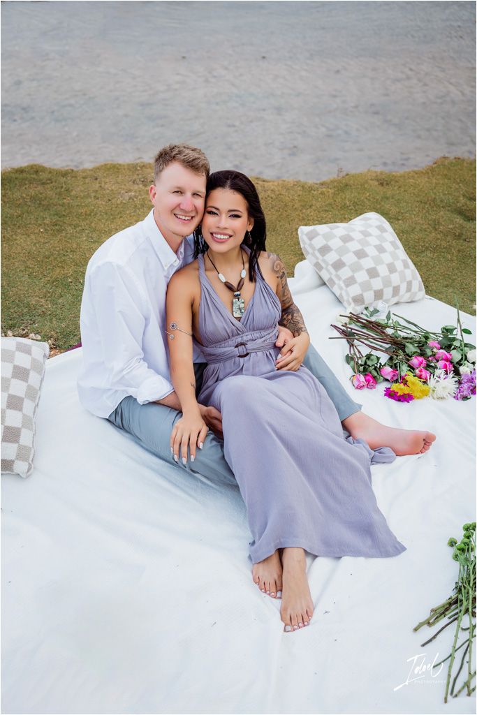 Couple enjoys romantic beach picnic on white blanket with flowers and decorative pillows.
