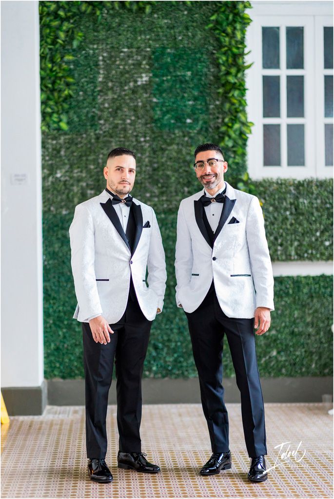 Two people wearing matching white tuxedo jackets with black lapels stand against an ivy-covered wall at a formal event.