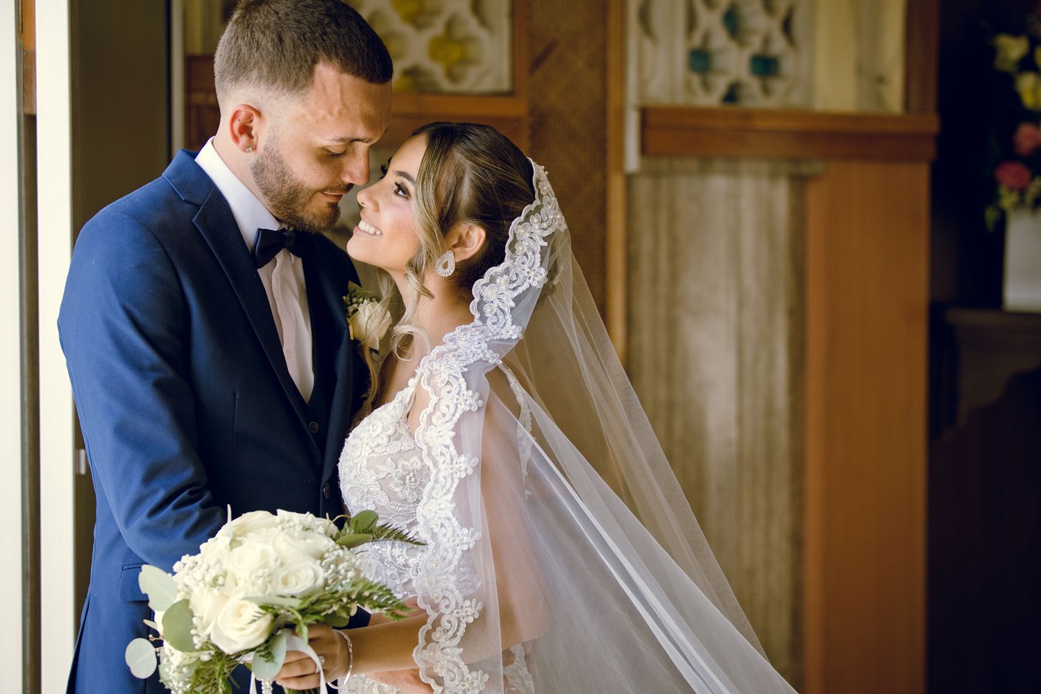 Bride and groom share a romantic moment, bride holding white bouquet in lace wedding dress with veil.