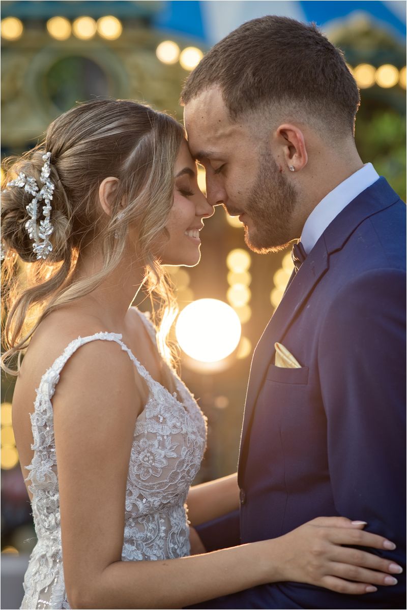 Bride and groom in romantic pose, foreheads touching, wearing wedding attire with bokeh lights in background.