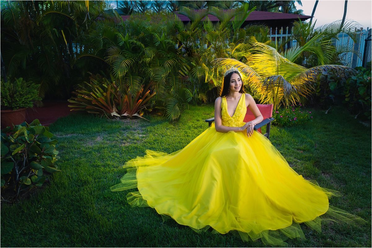 A bright yellow flowing dress twirls gracefully in a lush tropical garden setting with palm trees and greenery.