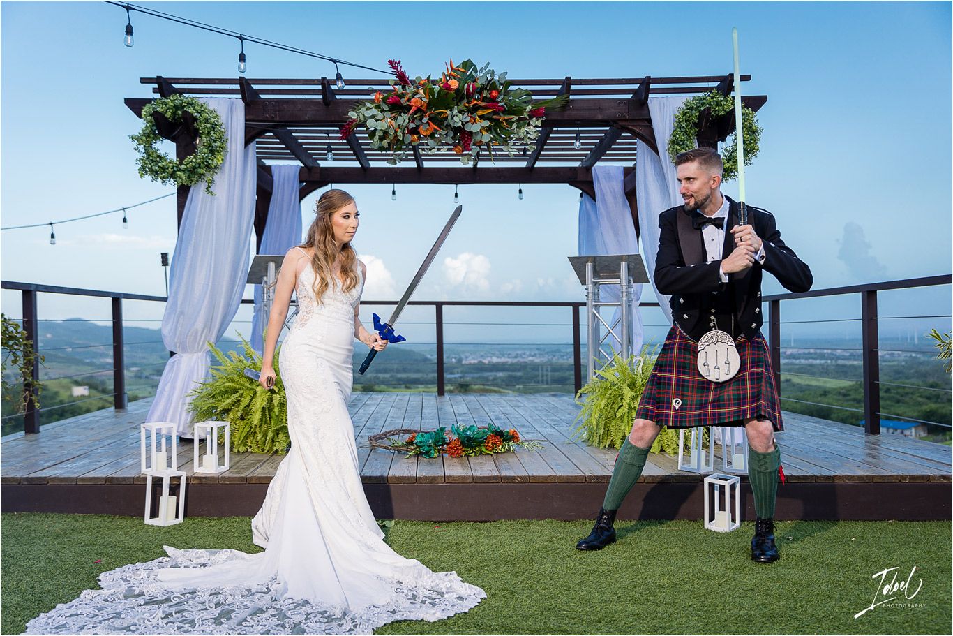 Couple performs playful sword fighting pose under decorated wedding pergola.