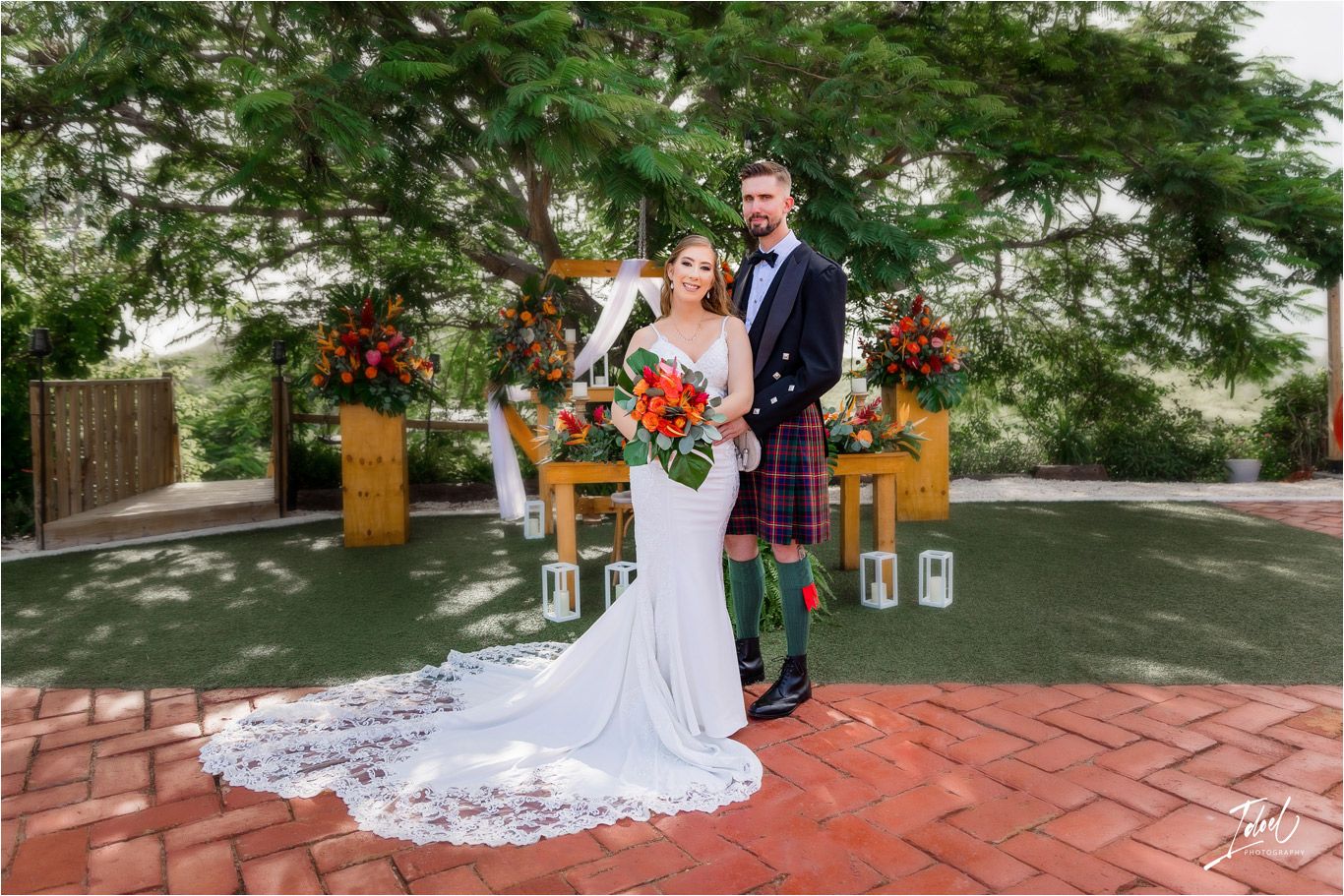 Couple in wedding attire pose together on brick patio with floral arrangements and Scottish elements.