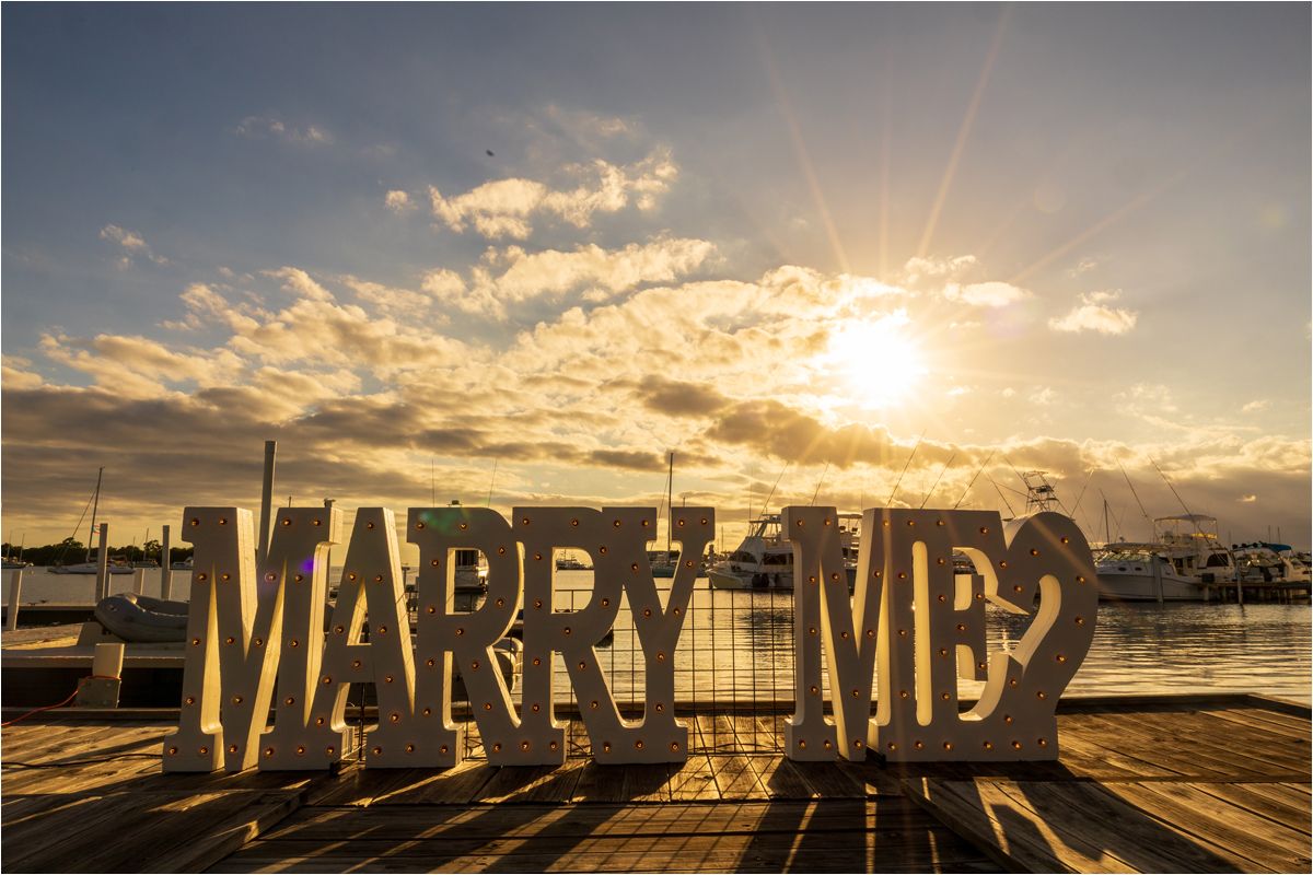 Large 'MARRY ME?' letters silhouetted against a dramatic sunset with sunbeams and clouds at a waterfront pier.