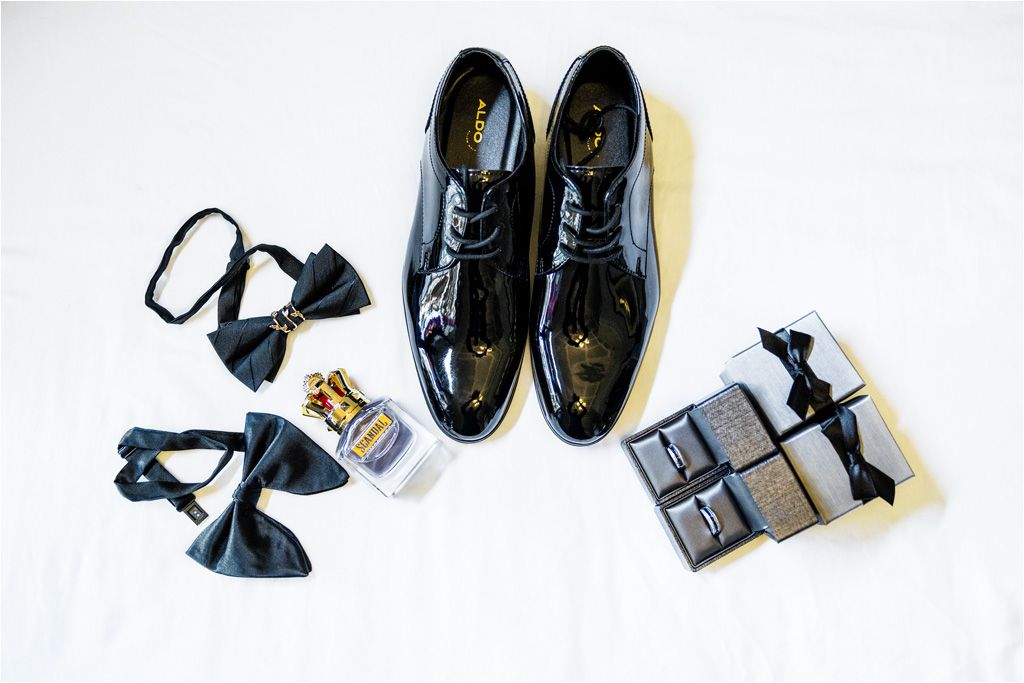 A flat lay of black formal leather shoes paired with dress socks, bow ties, and cologne on a white background.