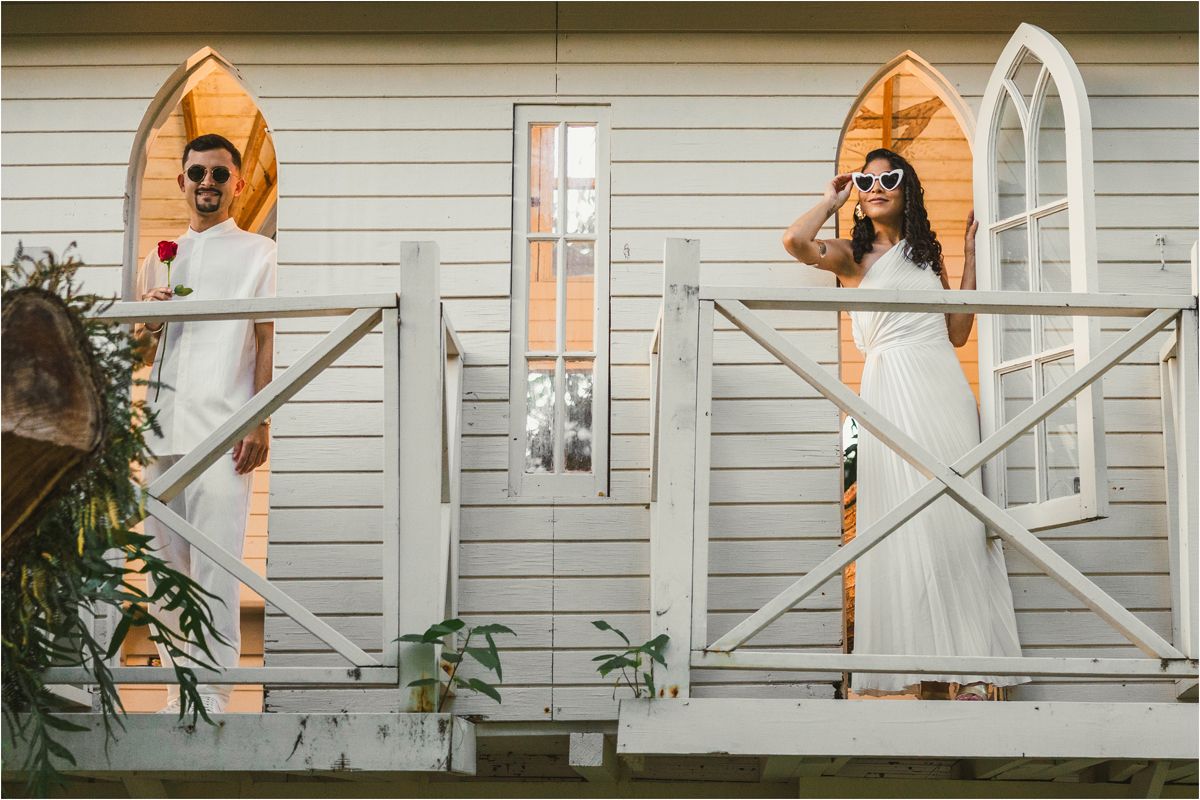 Two people in white dresses hold orange umbrellas while standing on a porch of a white house.